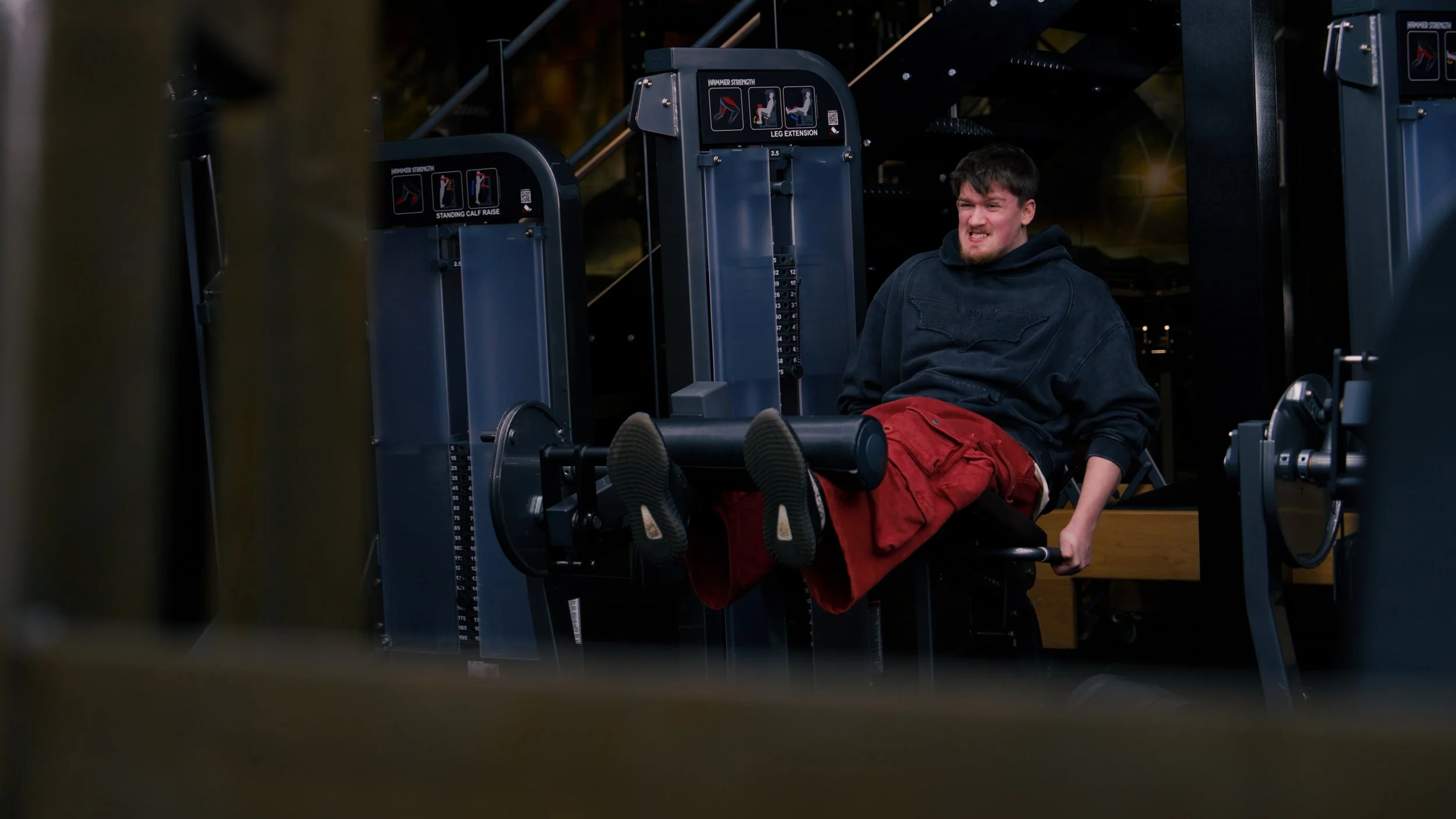 A young man in a black hoodie and red shorts lying on a leg press machine at a gym, grimacing while lifting weights.