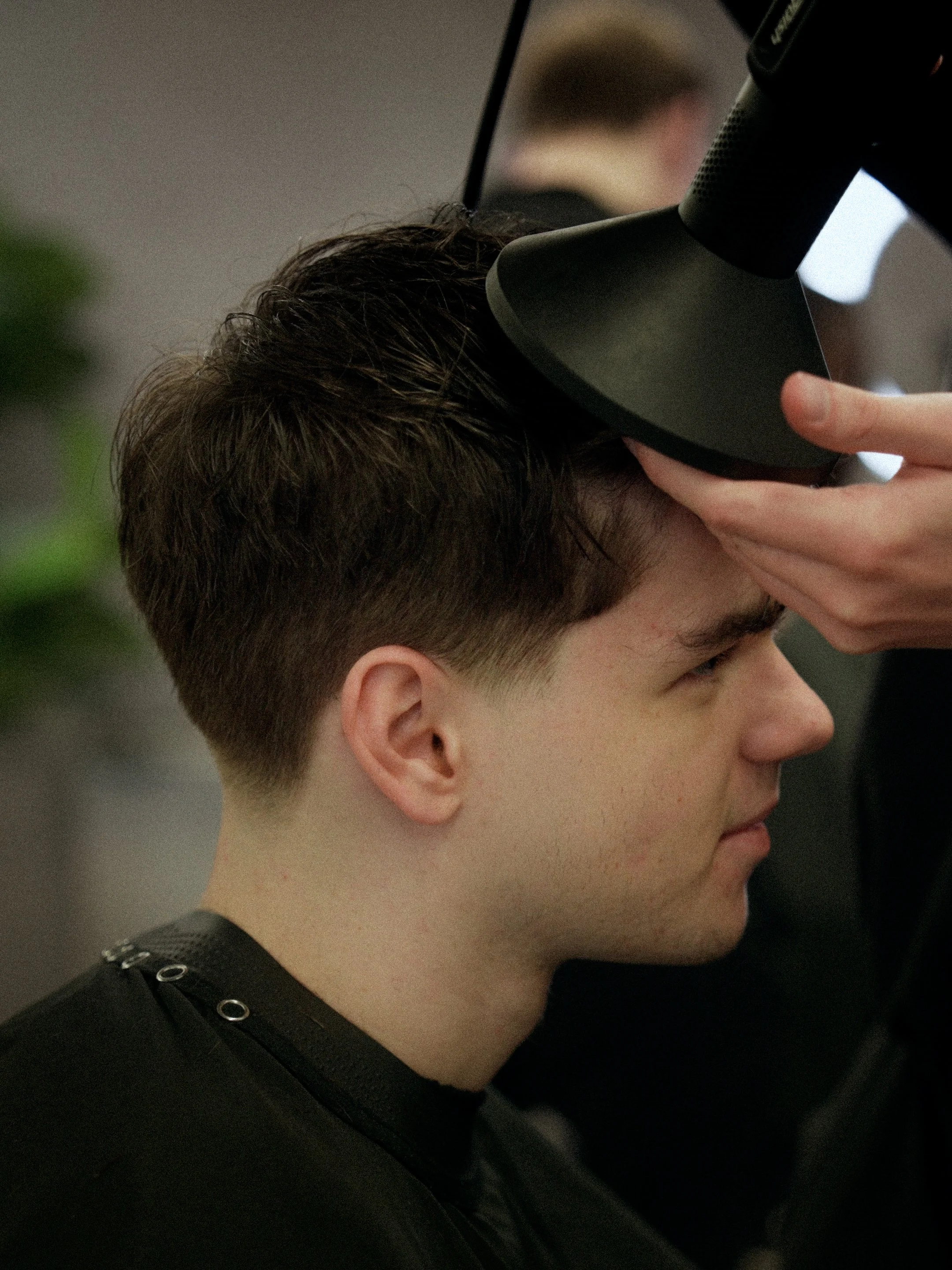A young man with brown hair getting a haircut at a salon, with a hairstylist using a hairdryer on his hair.