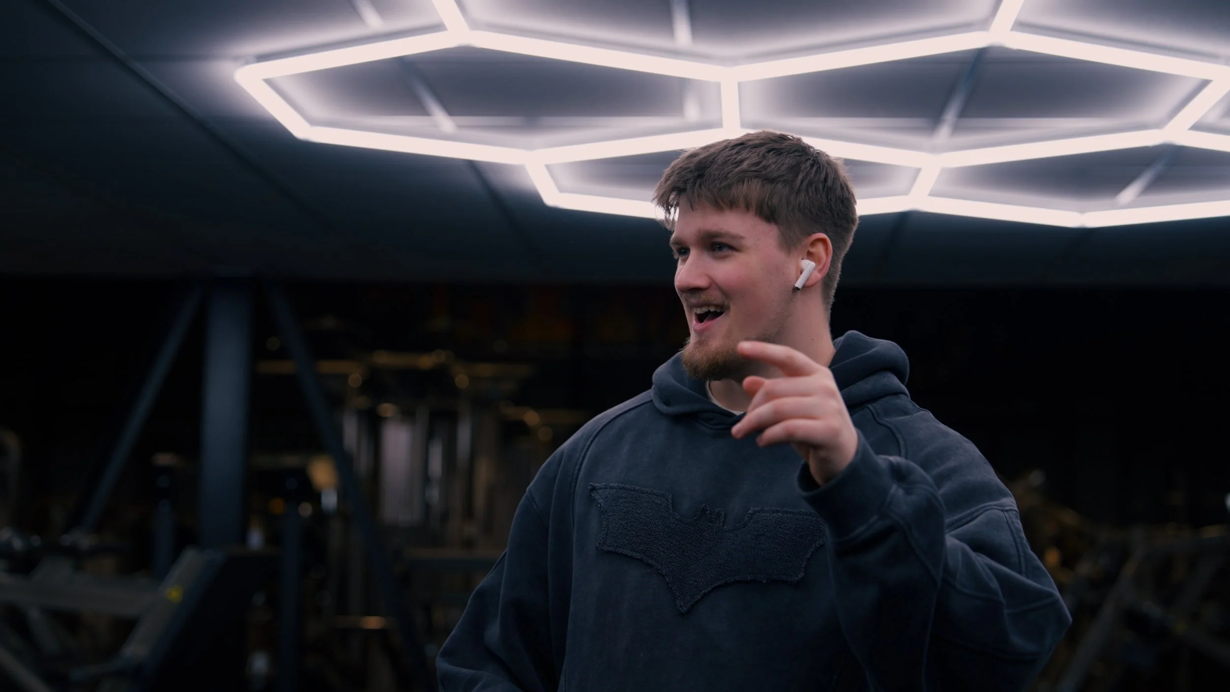 Young man with brown hair and a beard, wearing a black hoodie with a Batman logo, smiling and pointing, with white wireless earbuds, standing under illuminated hexagonal ceiling lights.