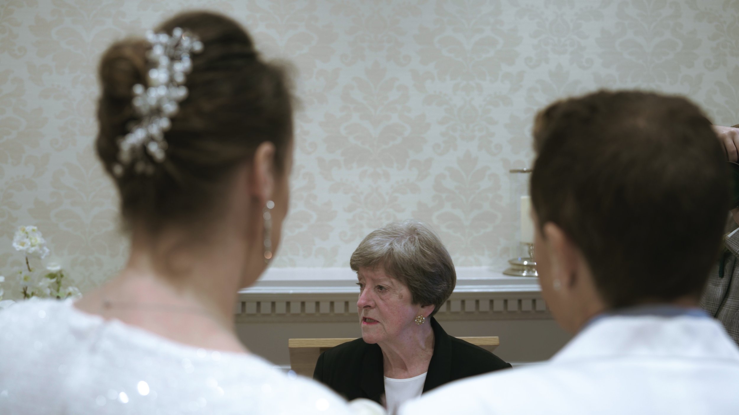 An older woman with short gray hair and a serious expression sitting at a table, with two women in wedding dresses in the foreground.