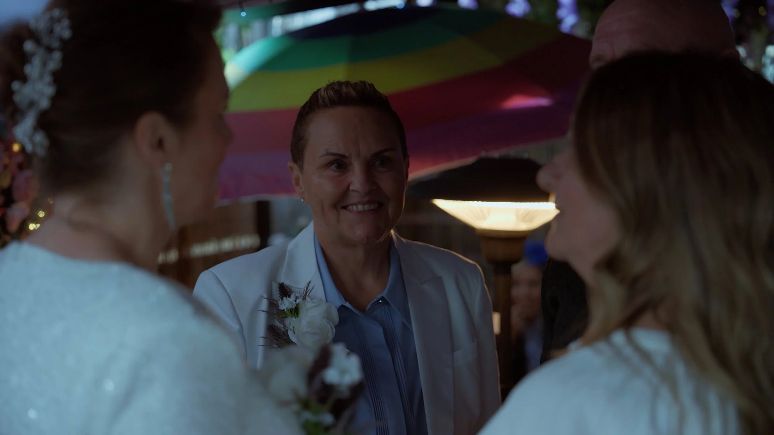 A woman with short hair and a white jacket smiling and talking to two women, one in a white wedding dress and the other with long hair, at a celebration under a multicolored umbrella.