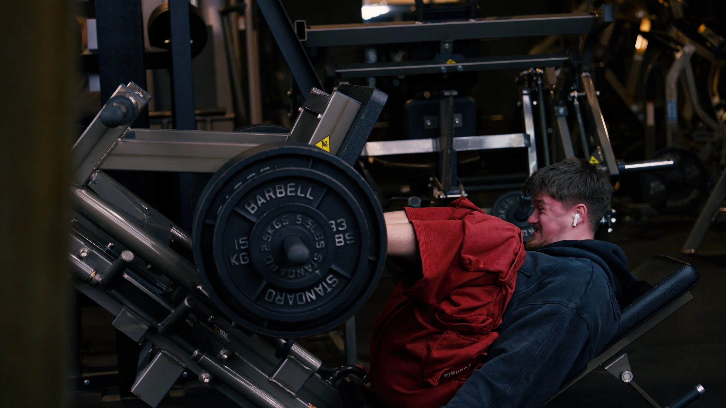 A man with dark hair and a beard, wearing headphones, smiling while lifting weights on a leg press machine at a gym.
