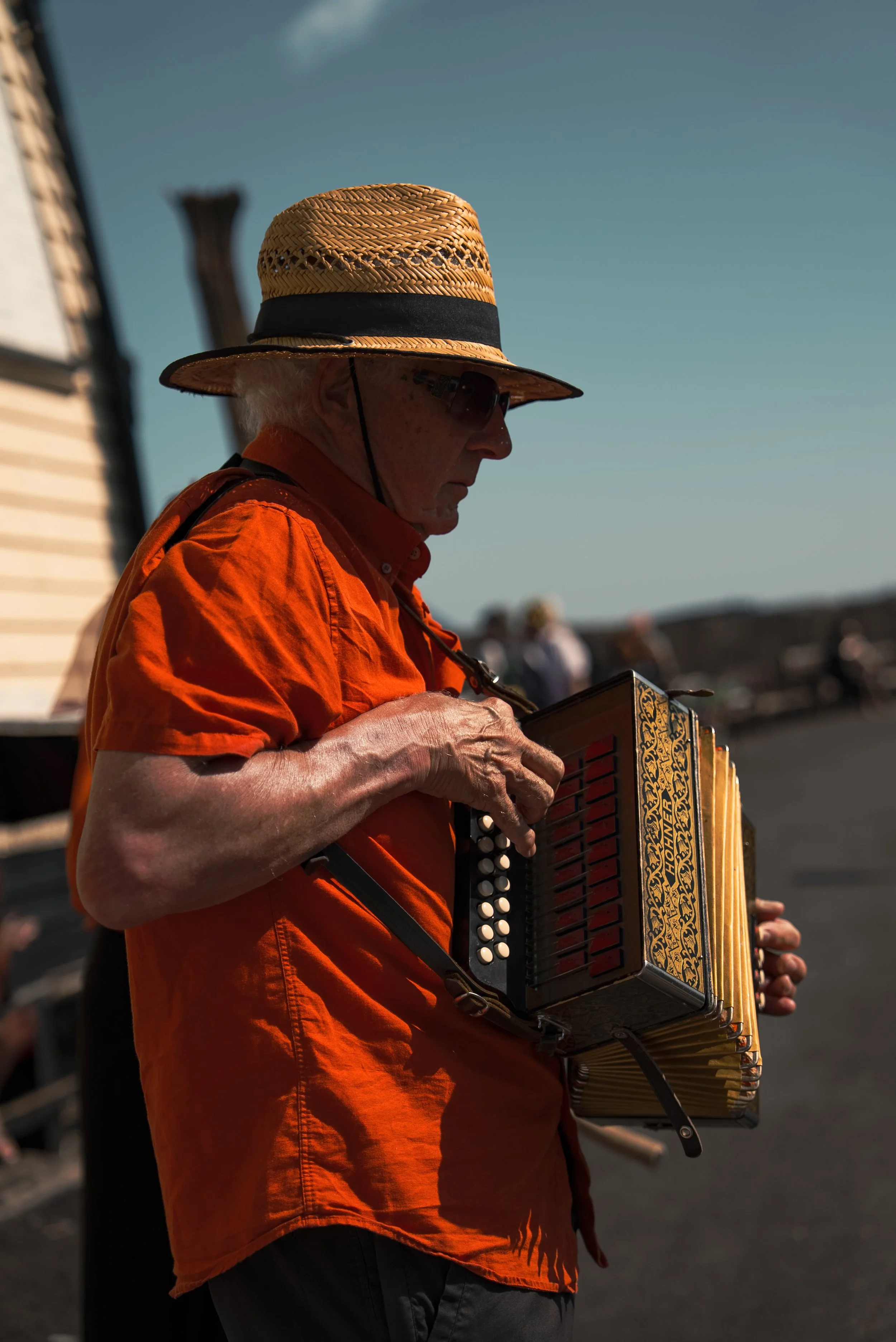 An elderly man wearing an orange shirt, wide-brimmed straw hat, and sunglasses playing a button accordion outdoors during daytime.