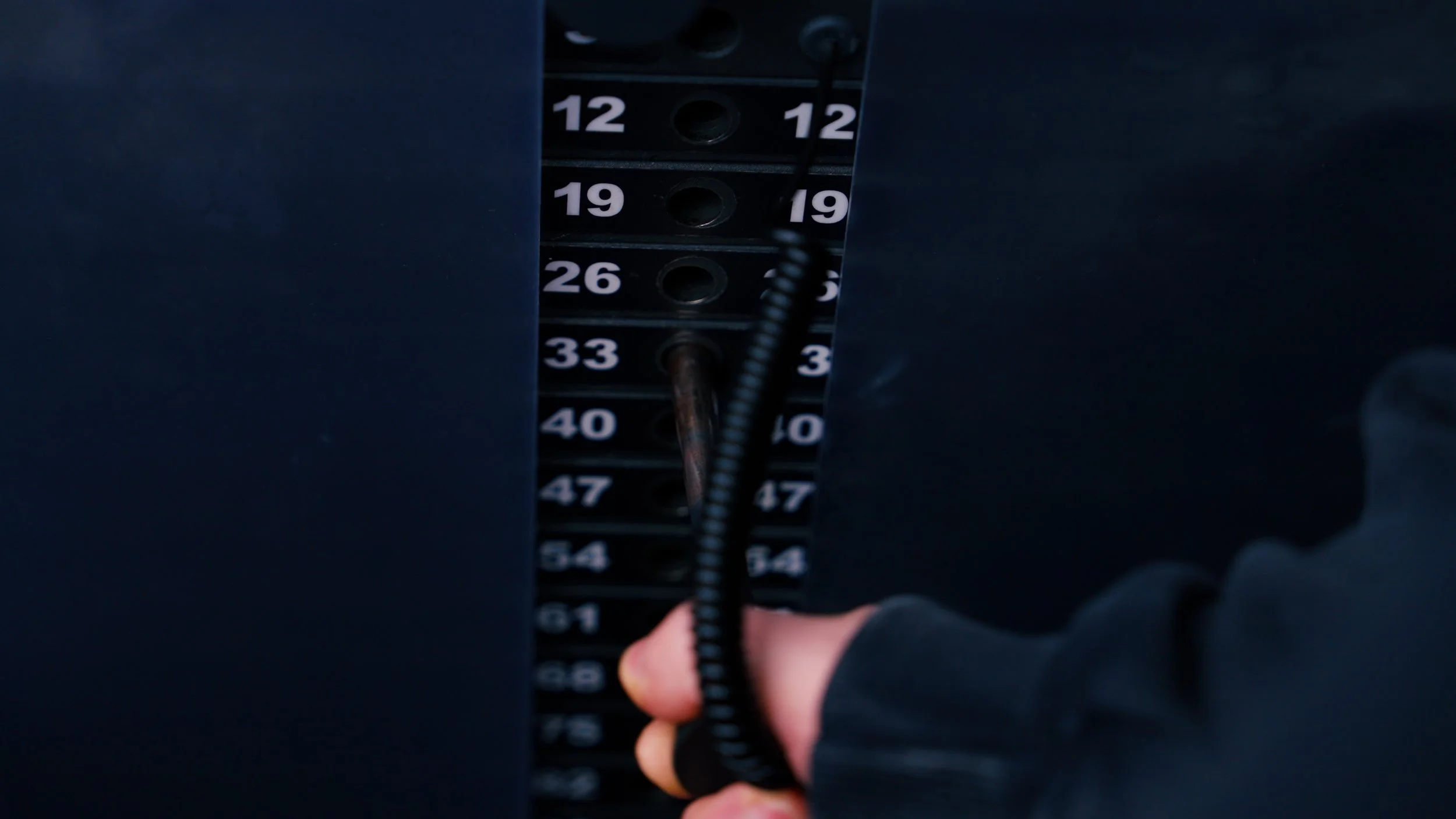 Close-up of a person's hand on an elevator button panel, with black panels numbered 12, 19, 26, 33, 40, 47, 54, and 61 visible. The person is pressing a button or about to press, and a black coiled cable is hanging near the panel.