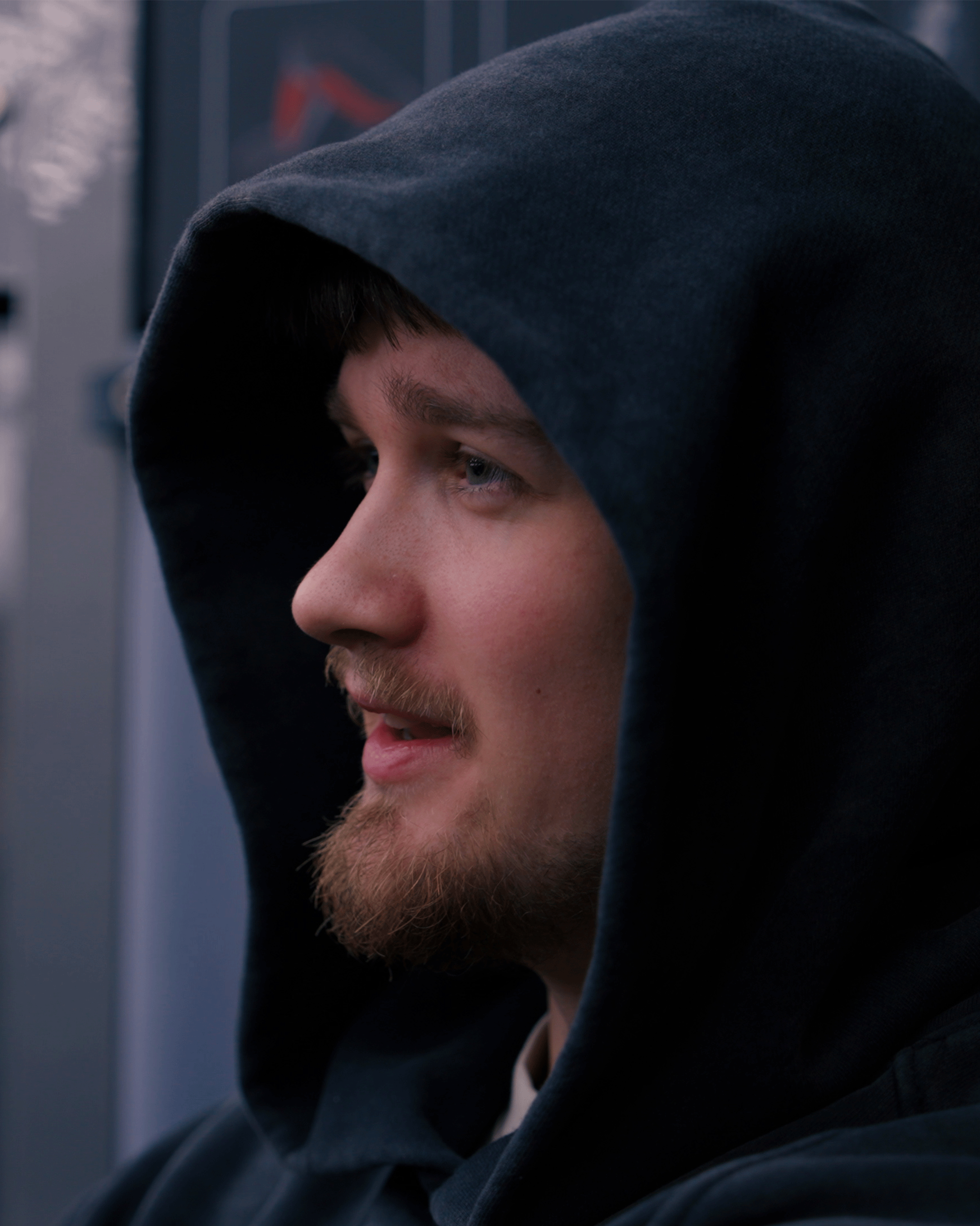 Close-up of a young man with a beard wearing a dark hoodie, sitting inside a vehicle.