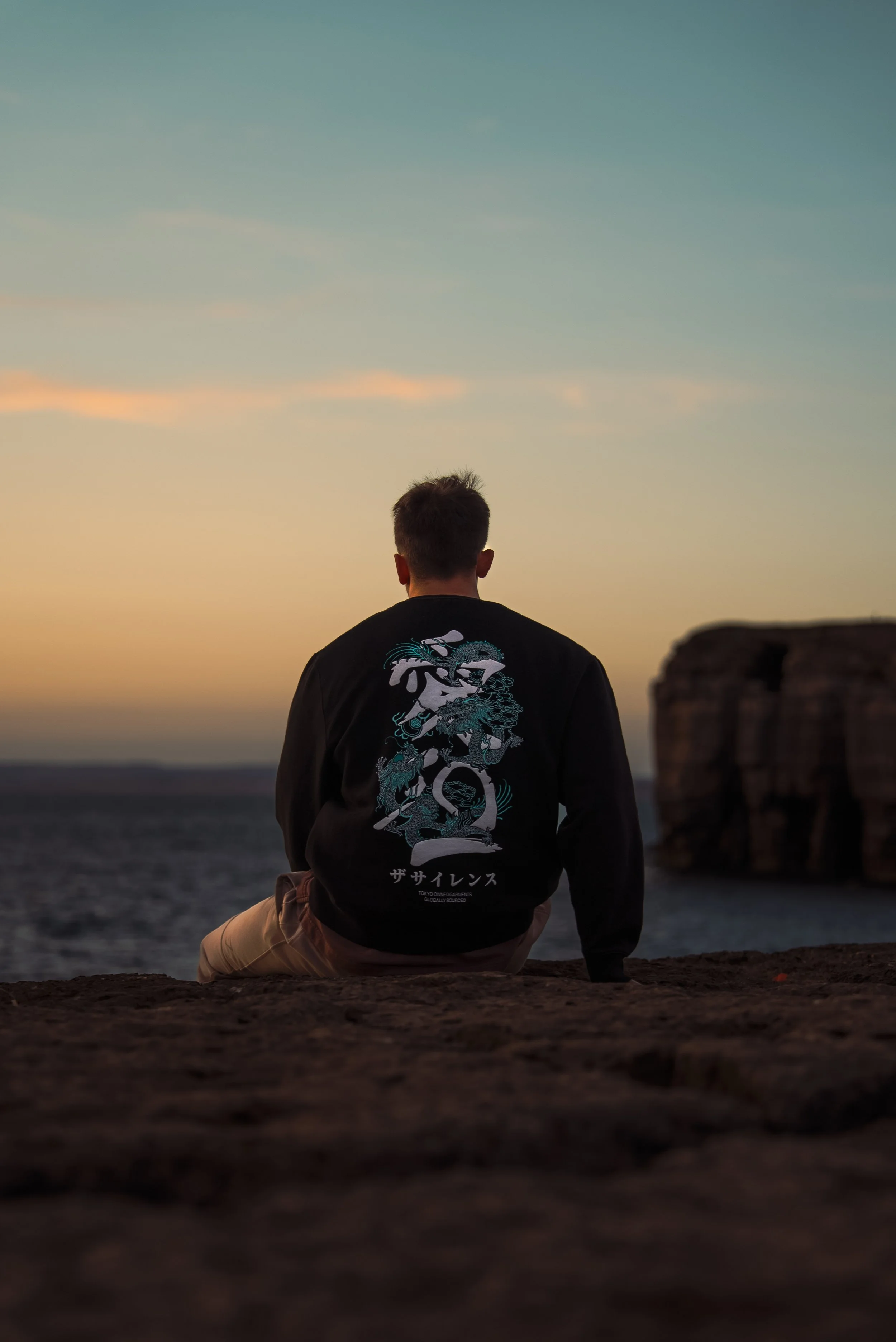A person sitting on a rocks overlooking the ocean at sunset, wearing a black jacket with a dragon design on the back.