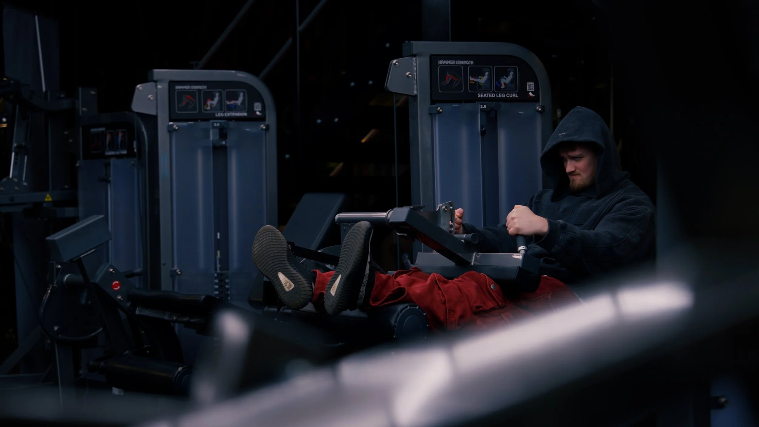 A man with a hoodie and red pants using a seated leg curl machine in a gym.