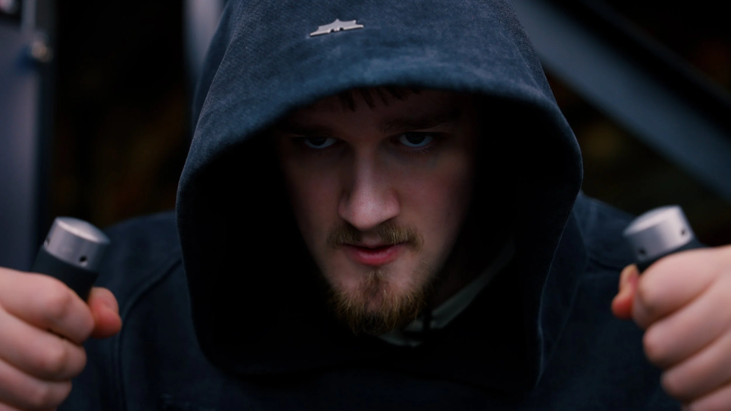A young man with a beard and blue eyes wearing a dark hoodie looks focused and intense while holding two dumbbells.