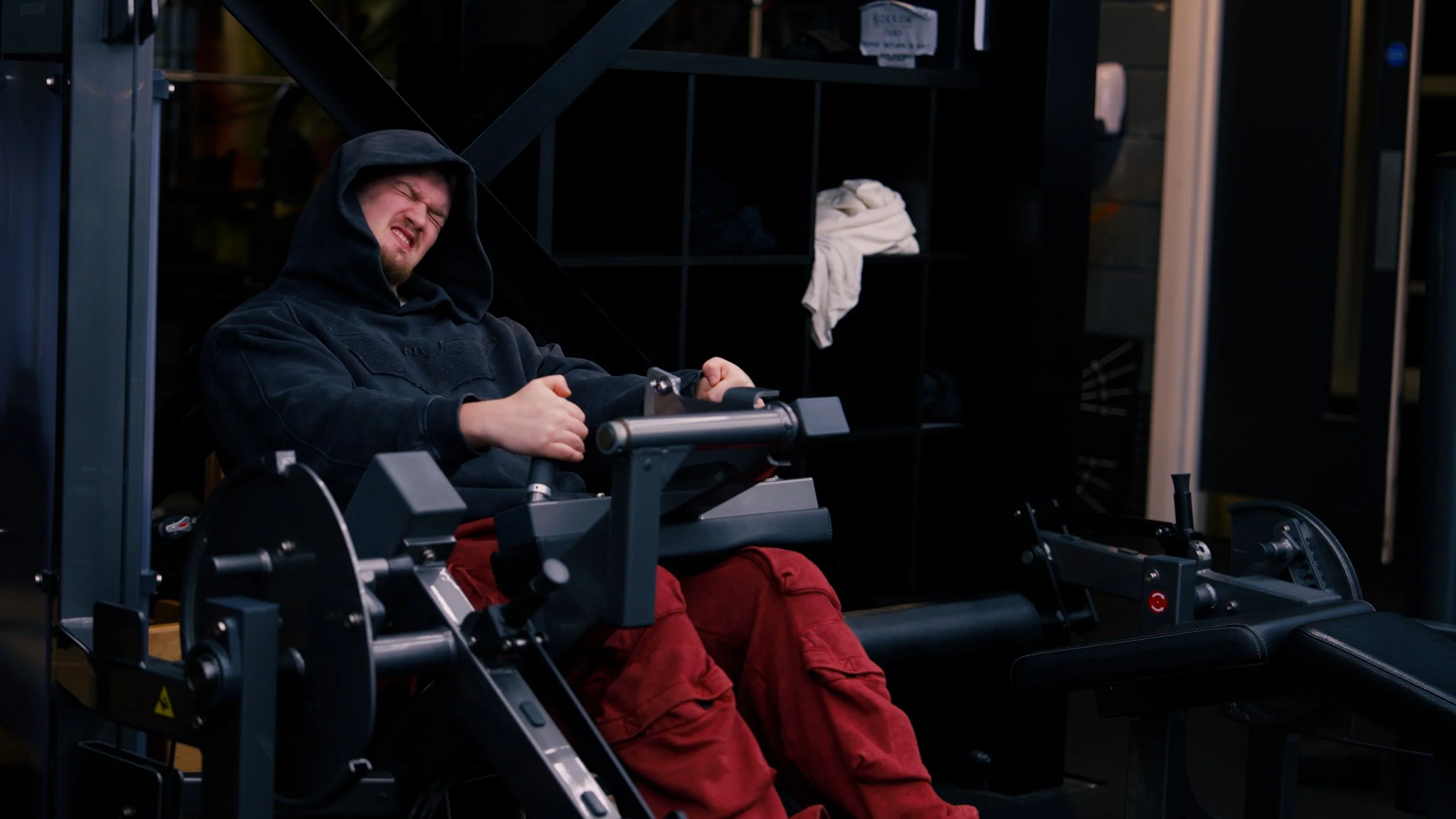 A man in a black hoodie and red pants is exercising on a rowing machine indoors, grimacing with effort.