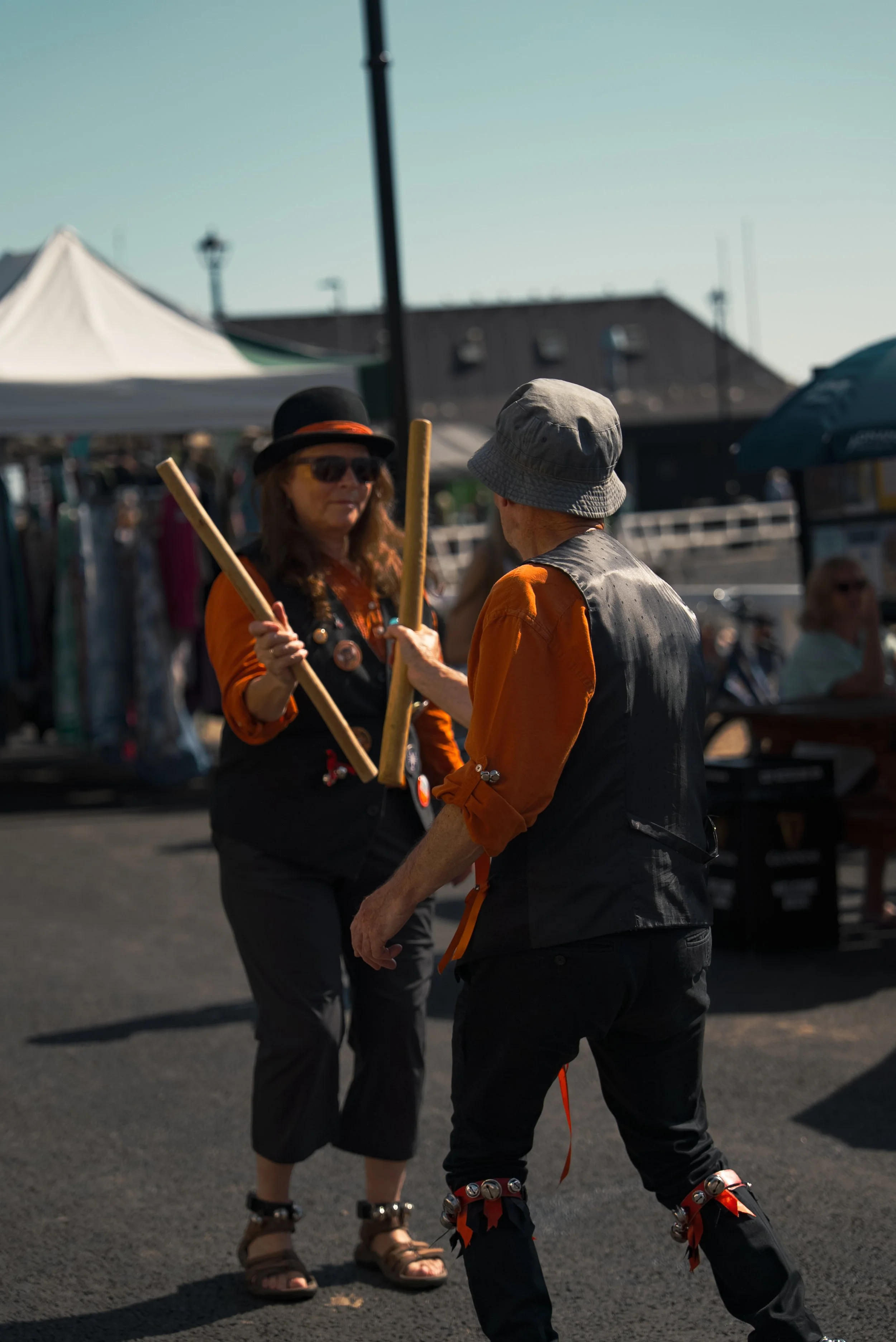 Two performers engage in a lively dance with wooden sticks at an outdoor festival or market, surrounded by tents and booths, under a clear blue sky.