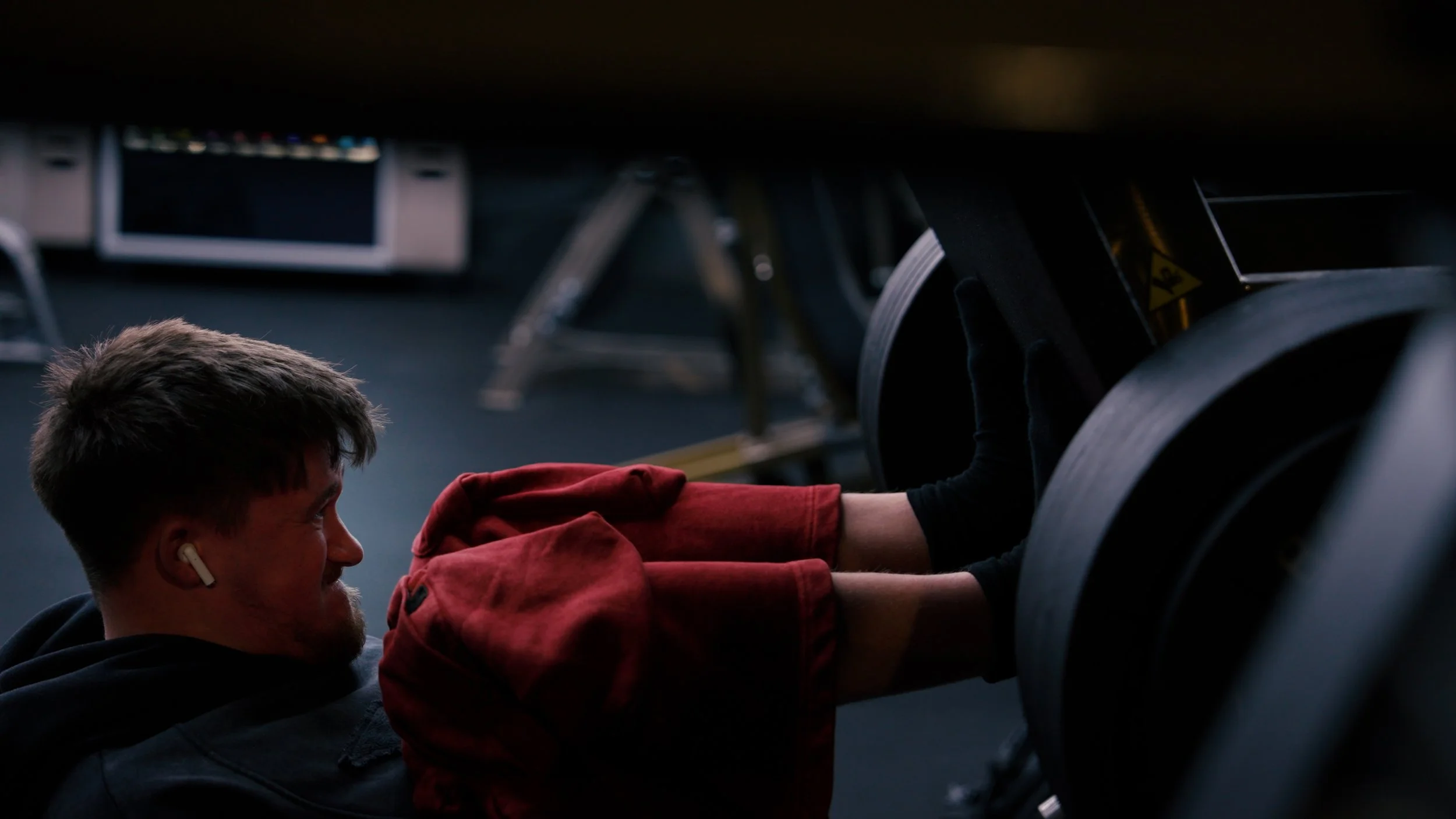 A man with airPods and a beard doing push-ups in a dimly lit gym, with weights and exercise equipment around.