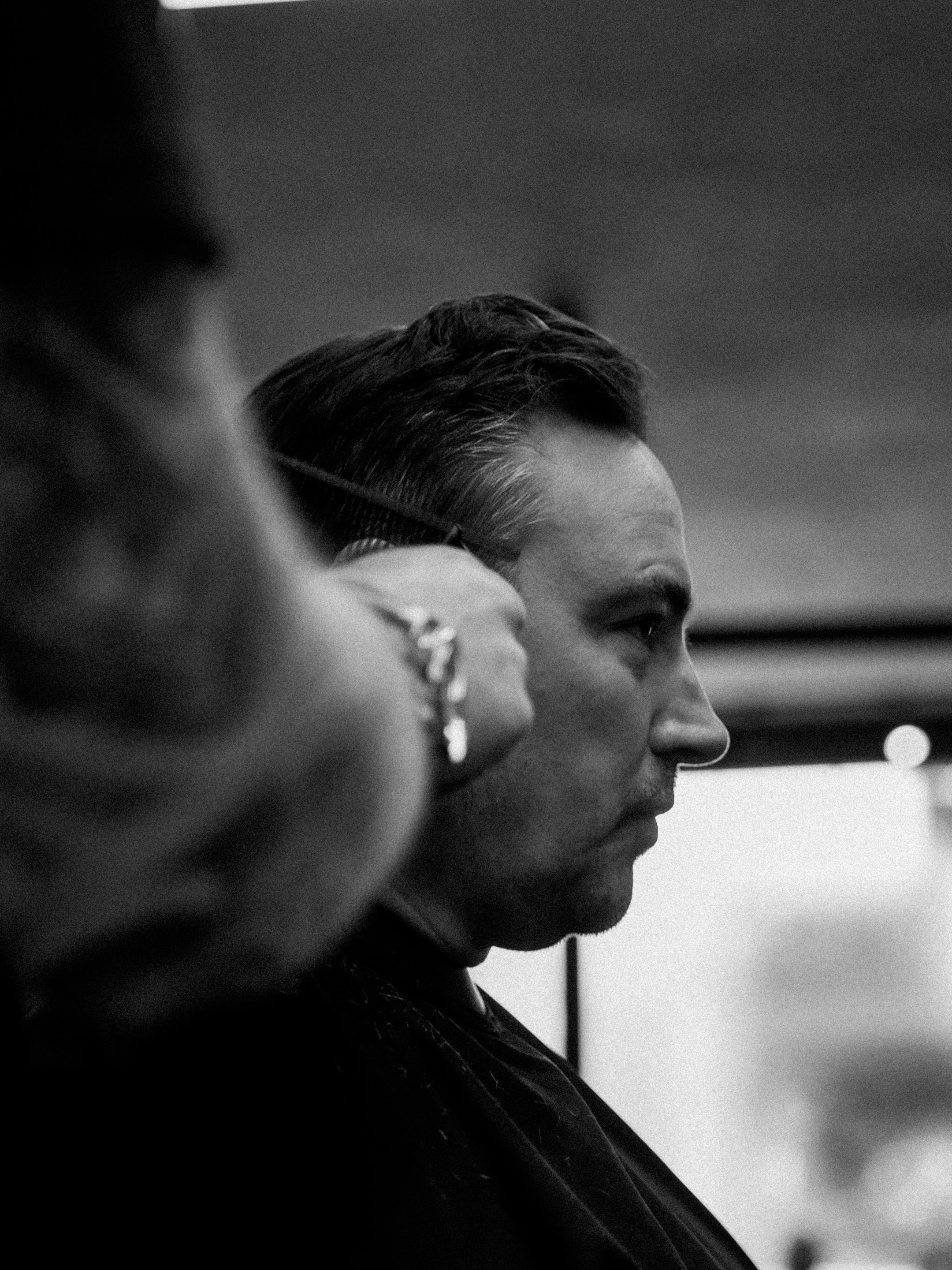 A black and white photo of a man with slicked-back hair getting a haircut, with another person partially visible in the foreground.