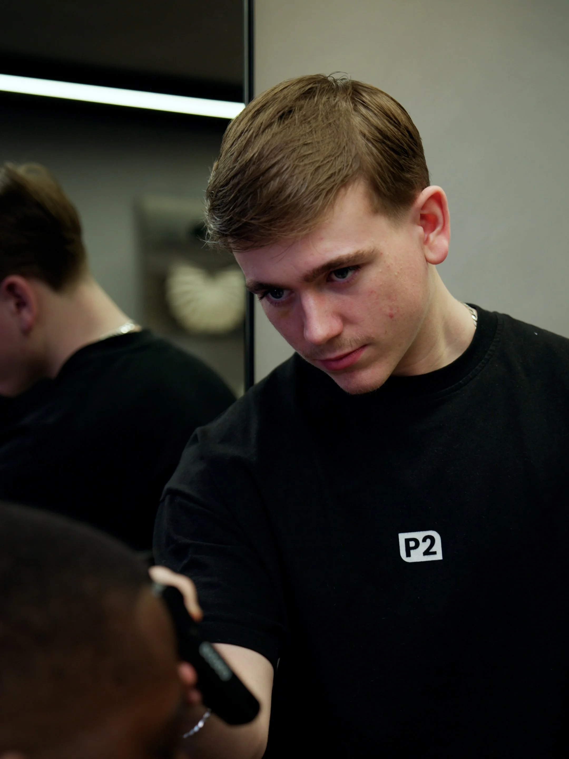 A young man with reddish-brown hair, wearing a black shirt with a white 'P2' on it, looks down at a person holding a microphone.