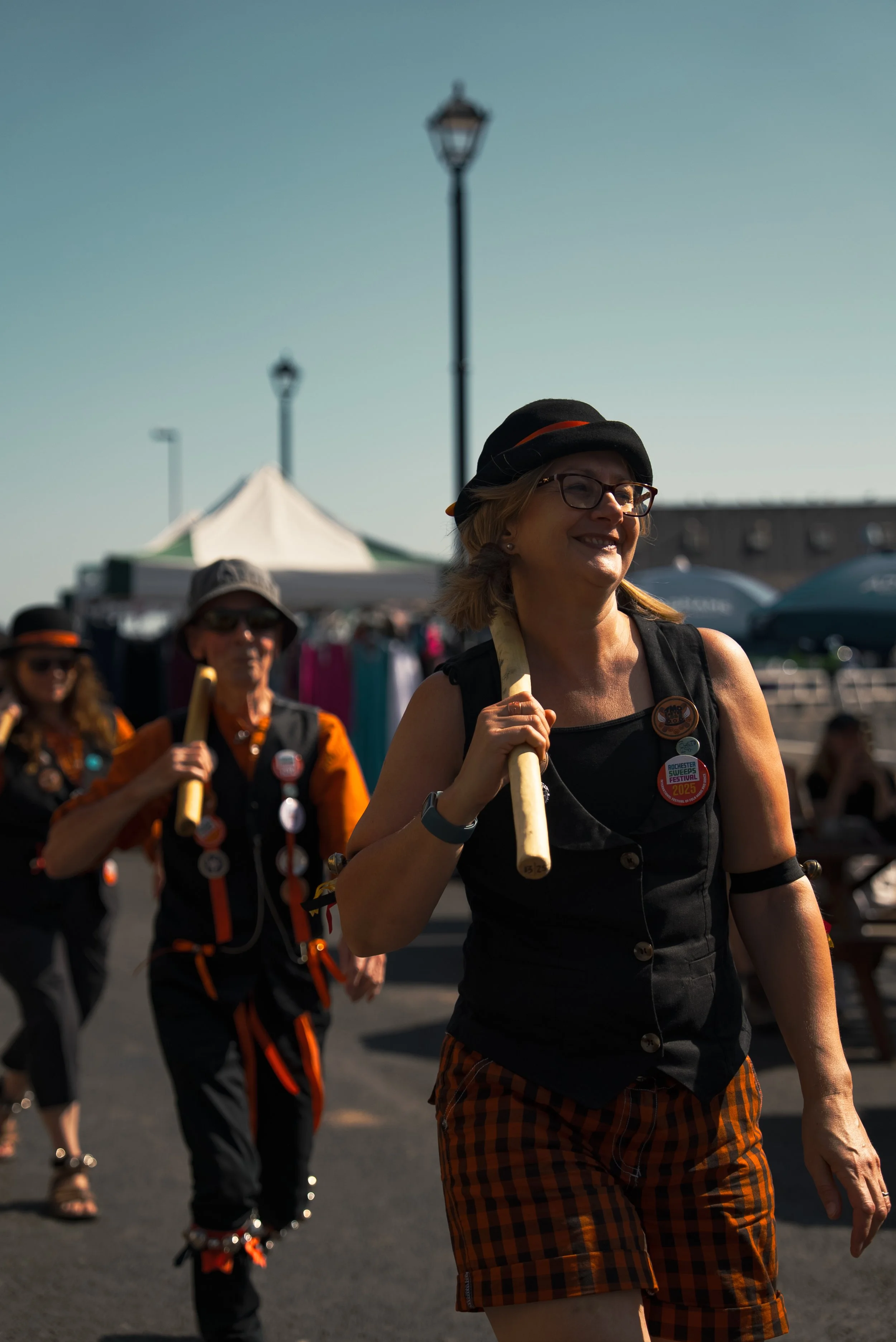 Women participating in a parade or festival, carrying wooden sticks, with tents and umbrellas in the background, sunny weather, casual and festive attire with badges and accessories.