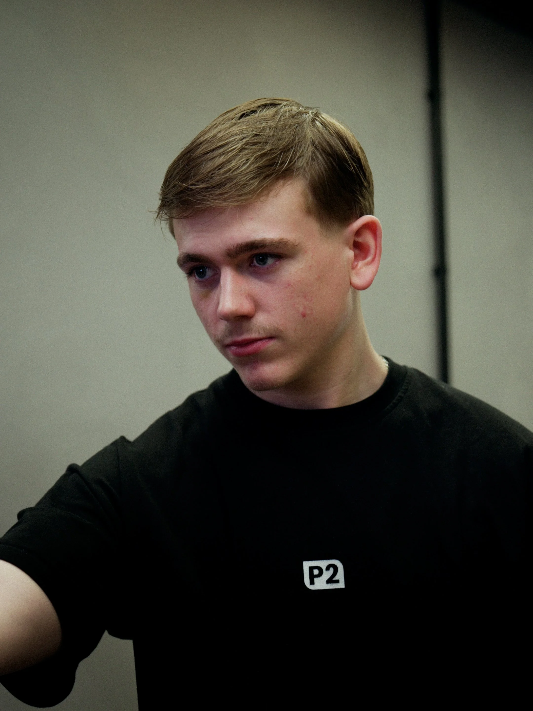 A young man with short blonde hair and blue eyes looking downward, wearing a black shirt with a white patch labeled 'P2'.