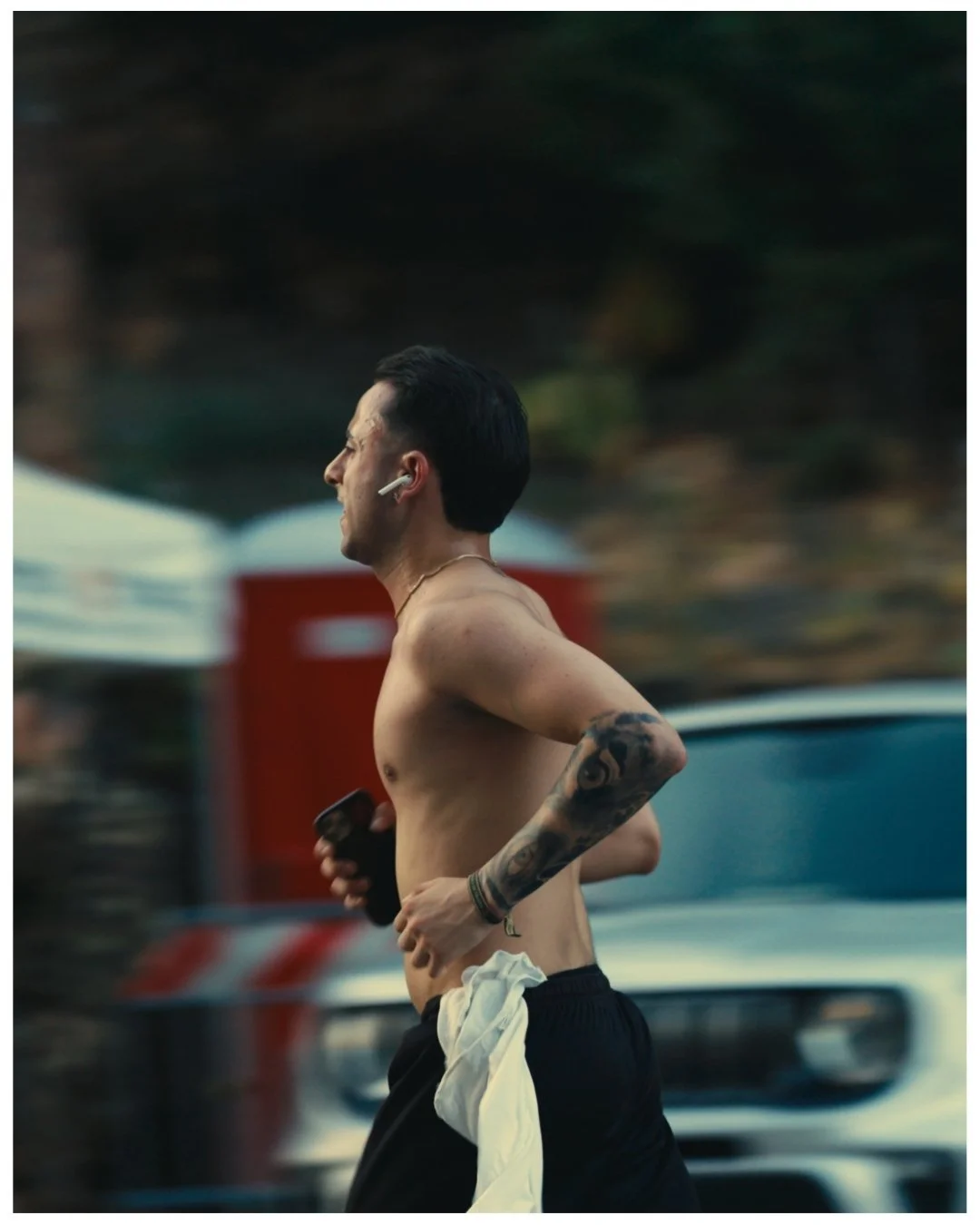 A shirtless man with tattoos on his left arm jogging outdoors, holding a smartphone in his right hand, wearing wireless earbuds, with a blurred background of vehicles and trees.