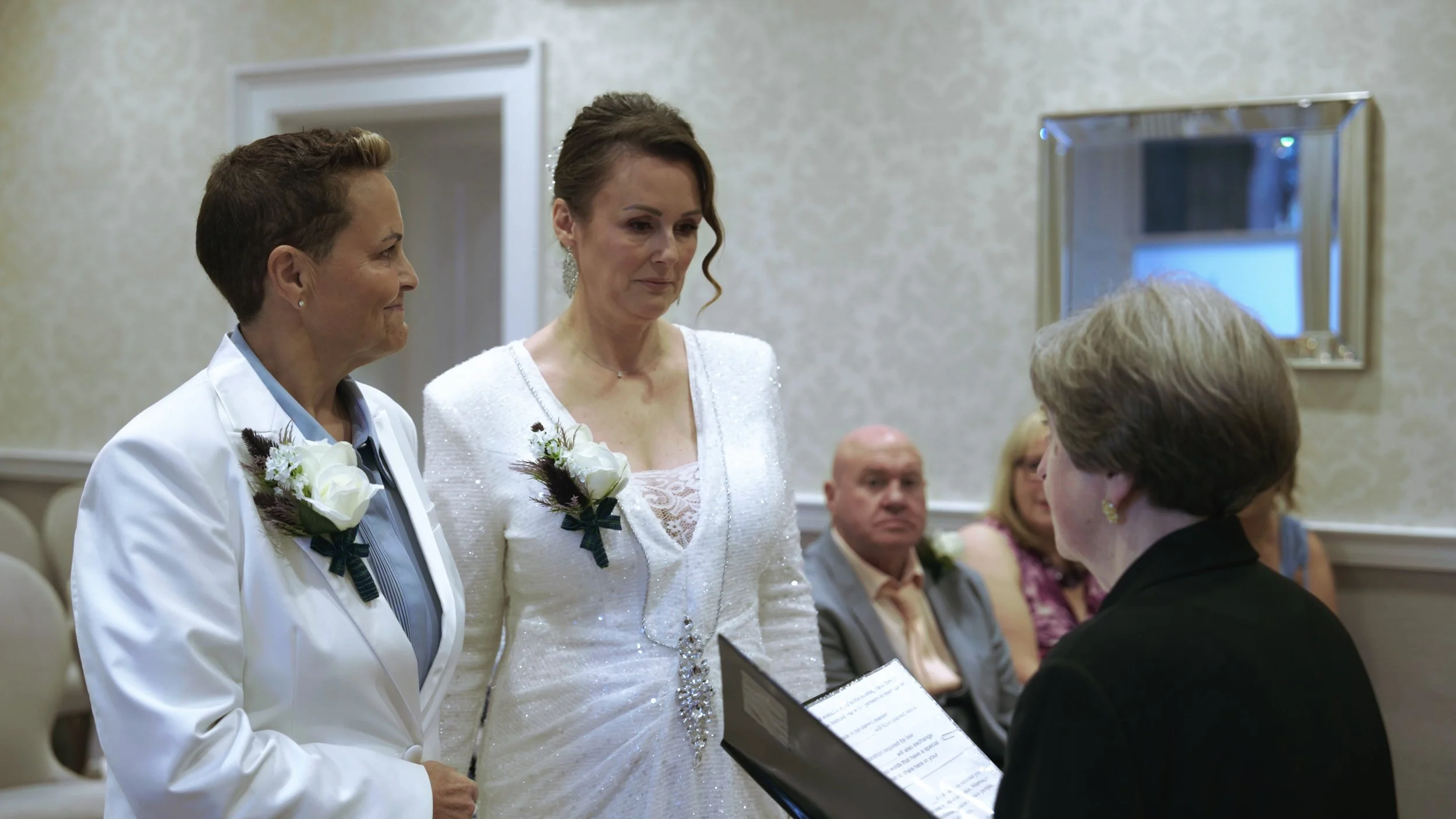 A wedding ceremony with a bride and groom standing before an officiant inside a decorated room with light-colored walls and a mirror on the wall.