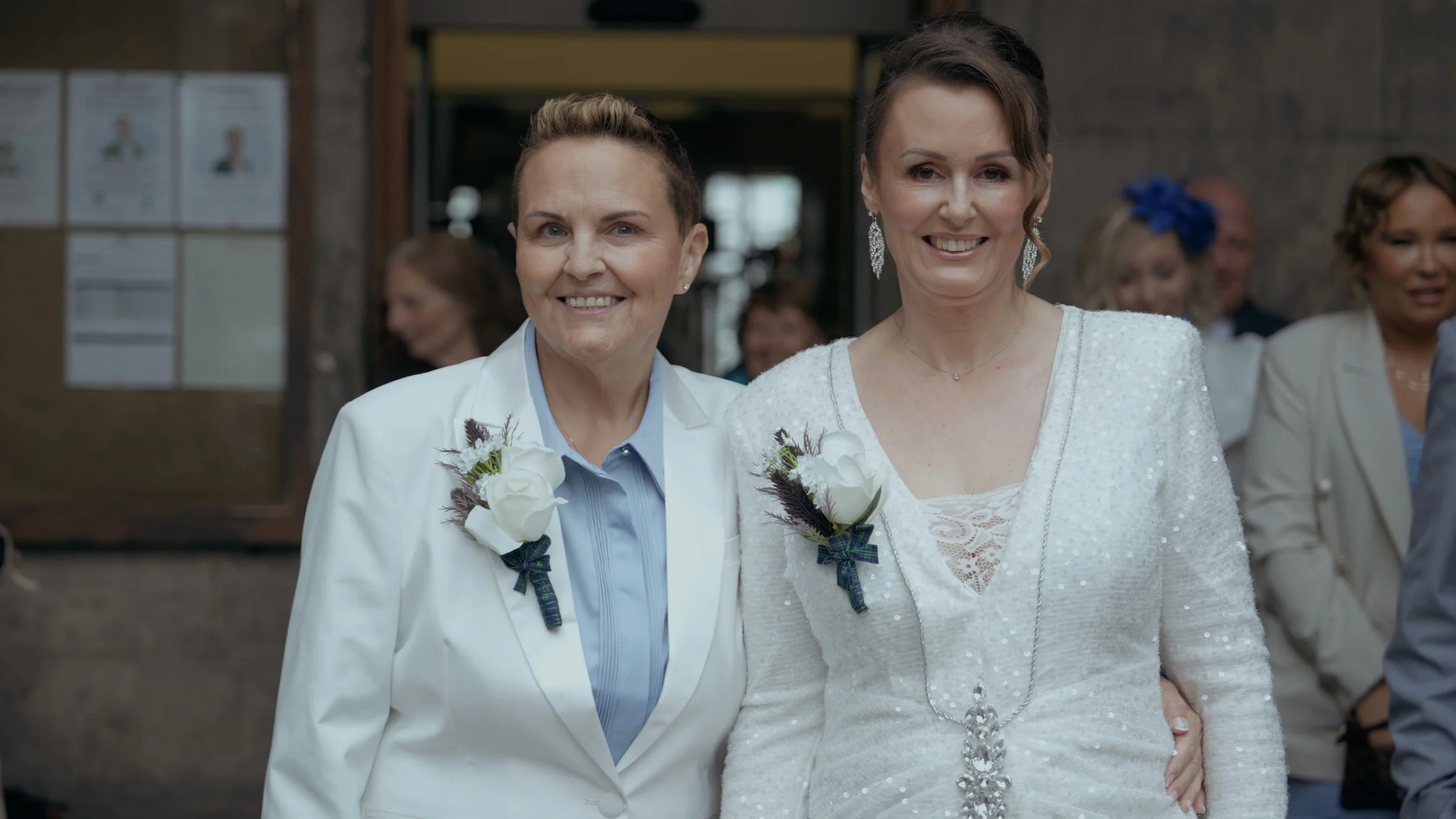 Two women at a wedding, smiling, with the woman on the right wearing a white dress and the woman on the left wearing a white suit. They both have corsages and are surrounded by guests.