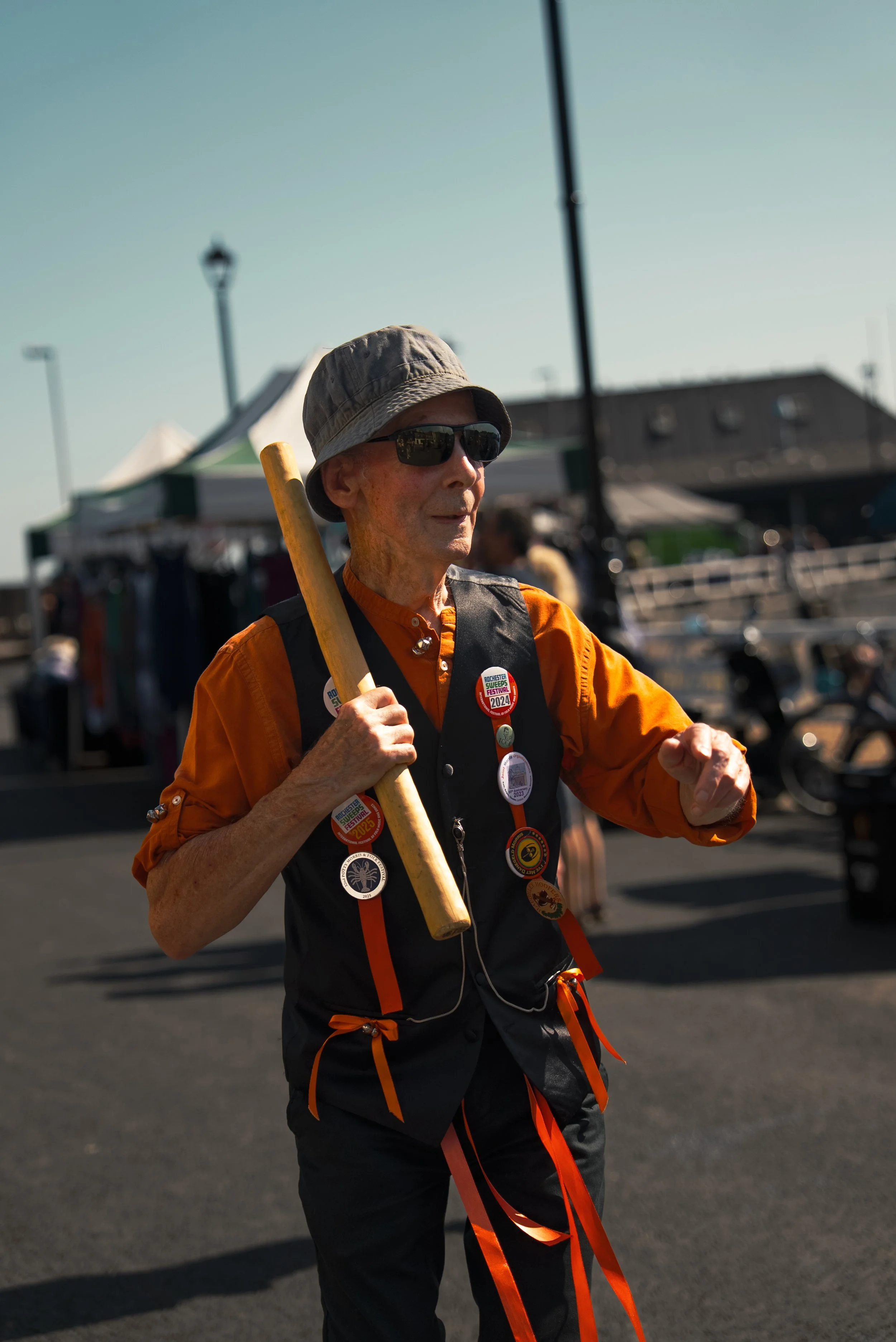 An elderly man wearing sunglasses, a gray hat, and an orange shirt, holding a wooden stick, standing outdoors during daytime at a bustling event with tents and people in the background.
