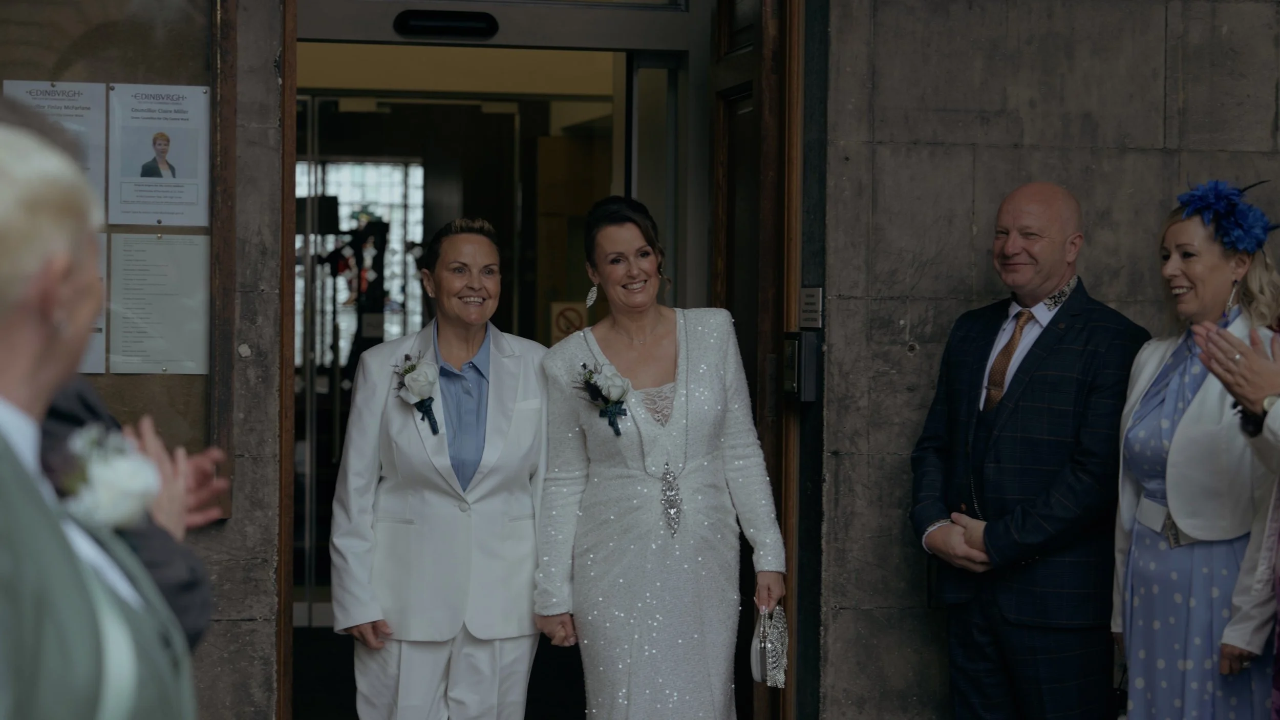Woman in a white sequin wedding dress holding hands with an older woman in a white suit, both smiling as they enter through an open door. Several people are on either side of the doorway, clapping and smiling, during a wedding celebration.