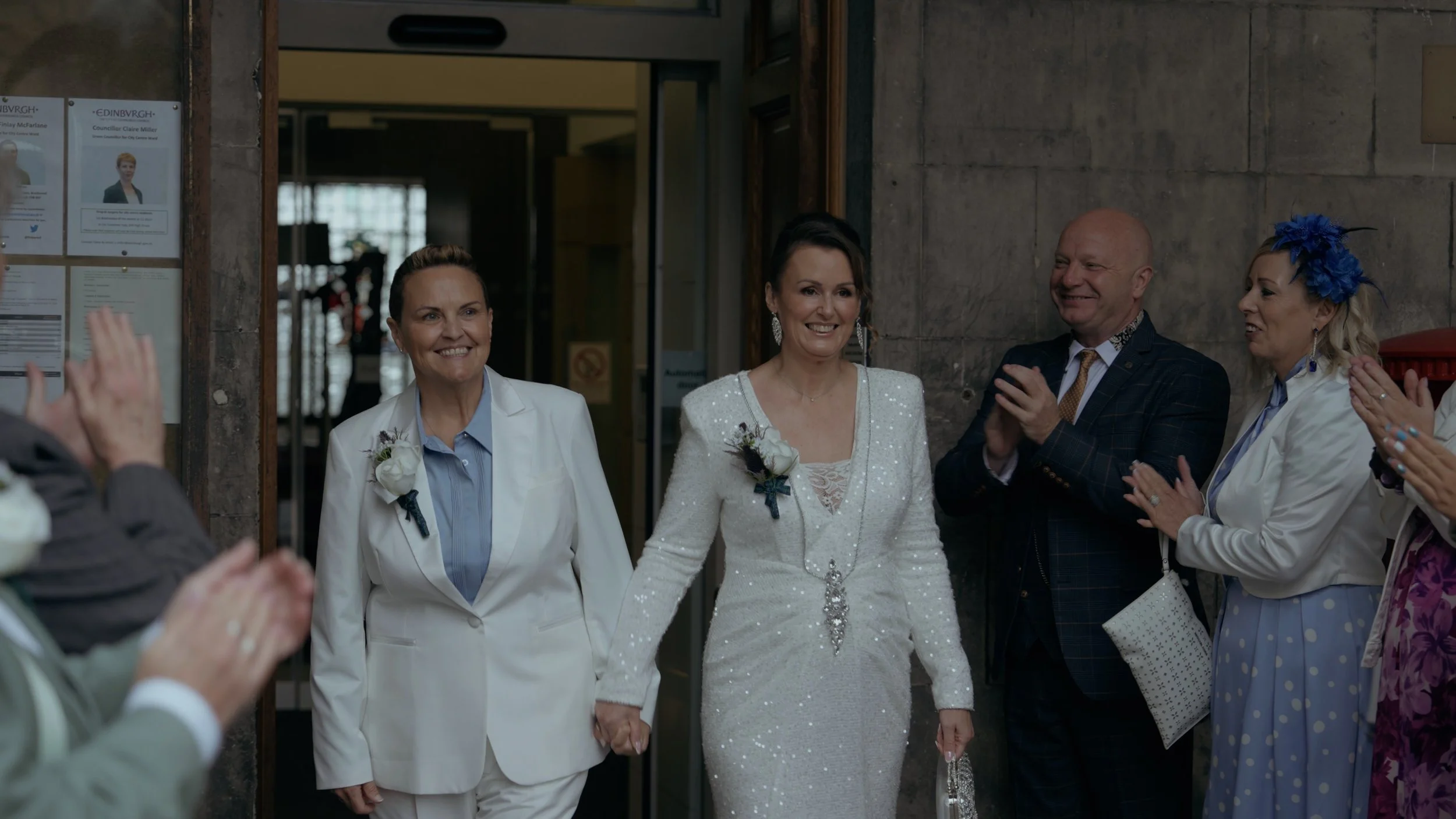 A bride in a white sequined gown holding hands with another woman in a white suit, walking into a room surrounded by clapping and smiling guests.