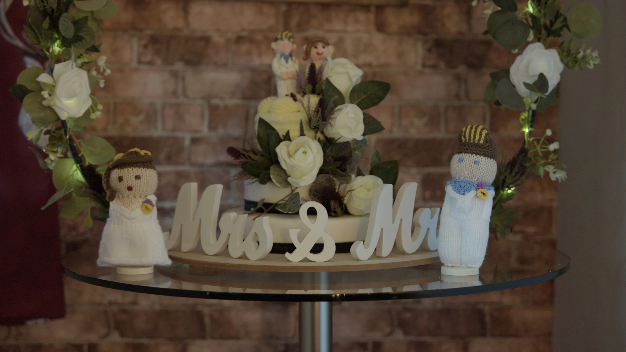 Wedding cake with white roses, two crocheted dolls dressed in white, and wooden "Mrs & Mr" signs on a glass table against a brick wall background.