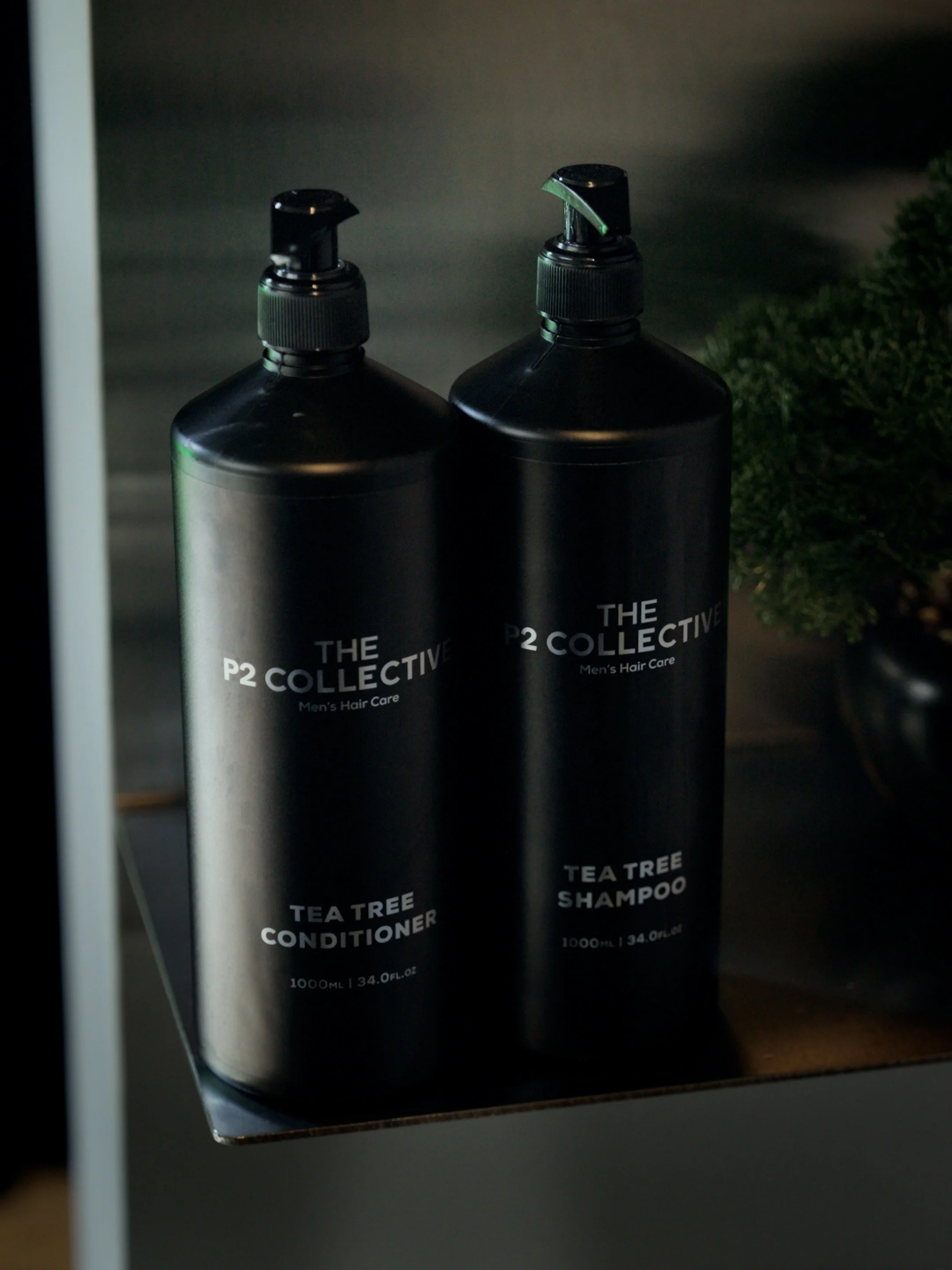Two black bottles of men's hair care products, labeled 'P2 Collective Tea Tree Conditioner' and 'P2 Collective Tea Tree Shampoo,' placed on a dark surface with some greenery nearby.
