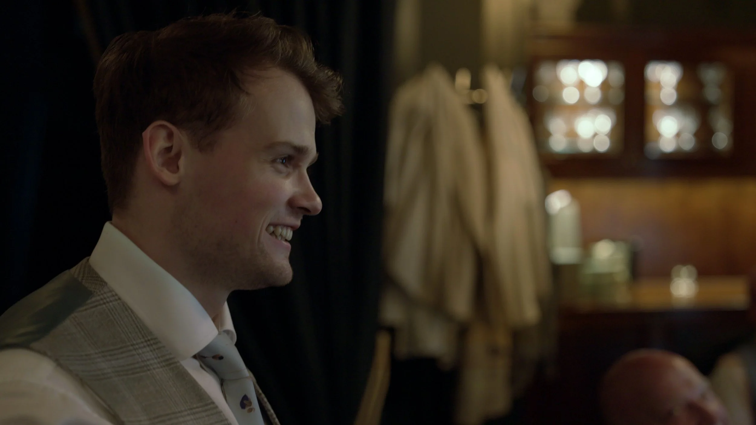 Close-up of a young man with light skin, smiling, in a dimly lit room, wearing a light-colored, plaid suit and a tie.
