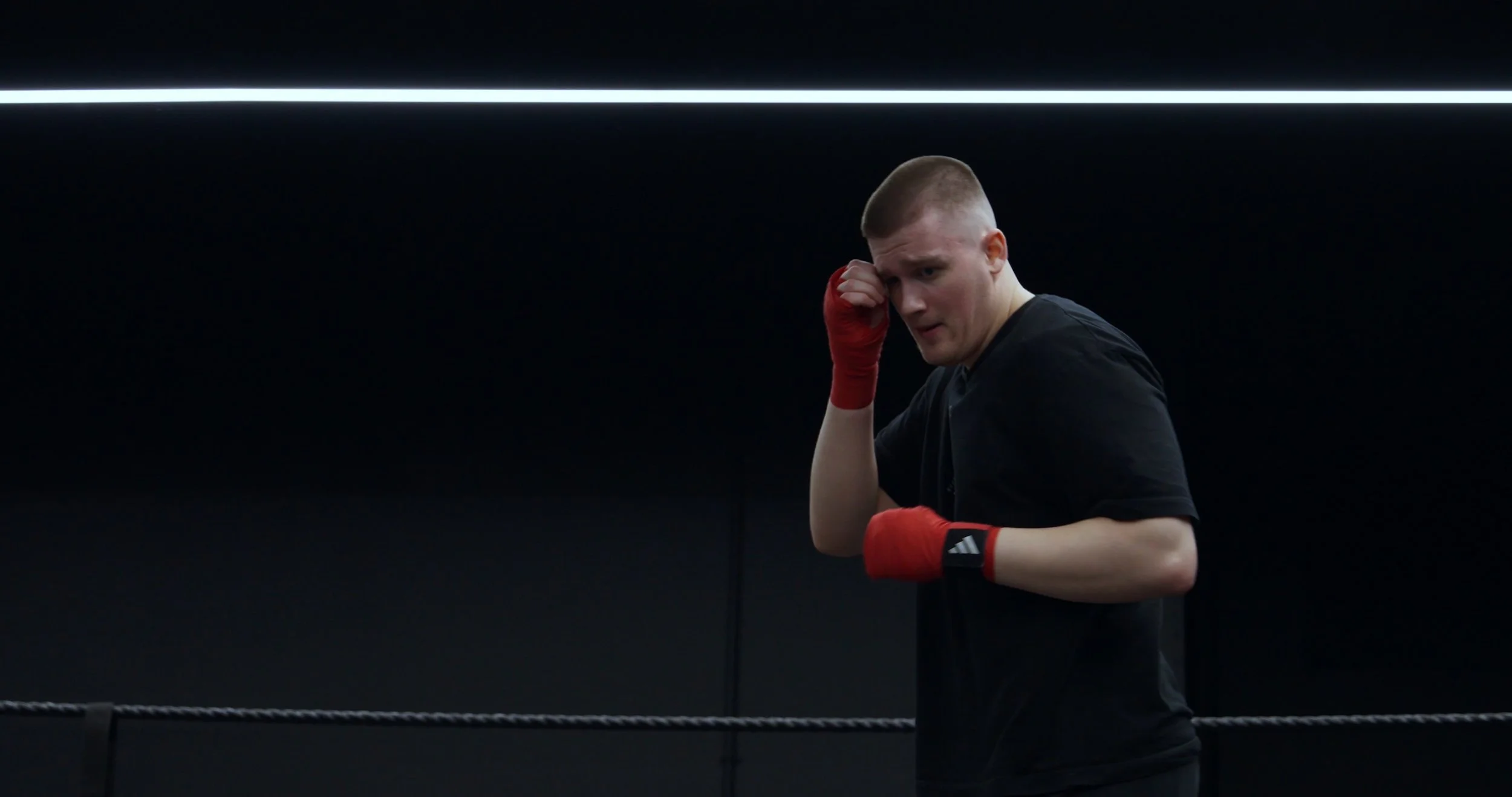 A young man in a black shirt with red boxing gloves holding his head in a gym or boxing ring.