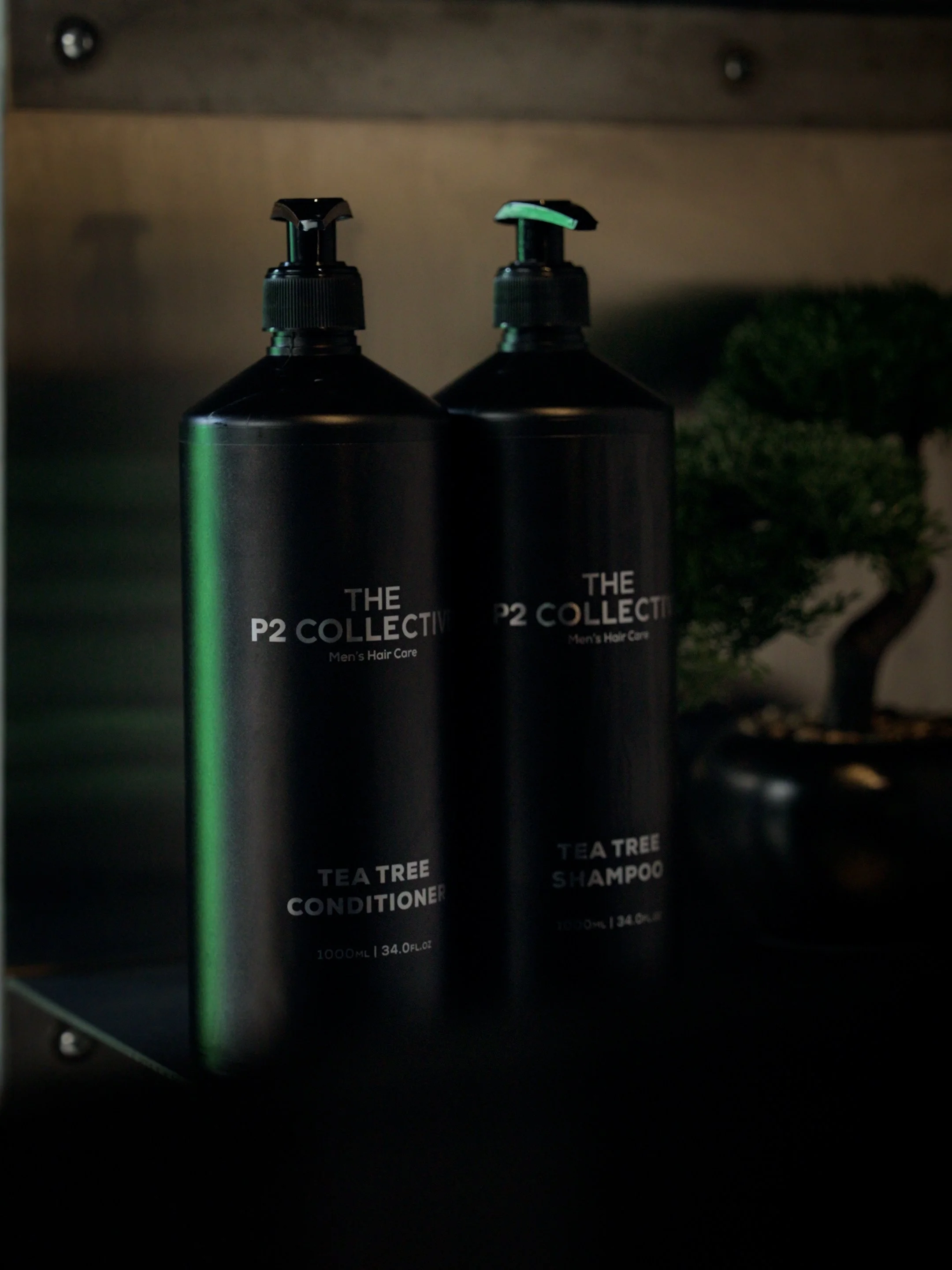 Two black bottles of men's hair care products, one labeled 'Tea Tree Conditioner' and the other labeled 'Tea Tree Shampoo', standing on a black shelf with a small plant and wooden background.