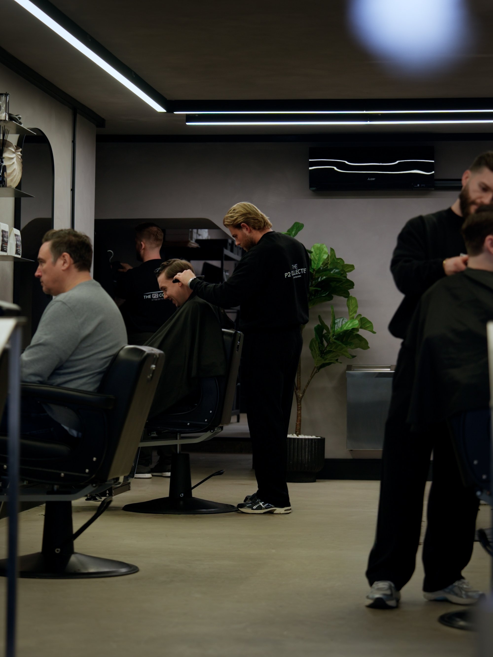 Barbers cutting men's hair in a modern salon with dark walls, a large plant, and minimal lighting.