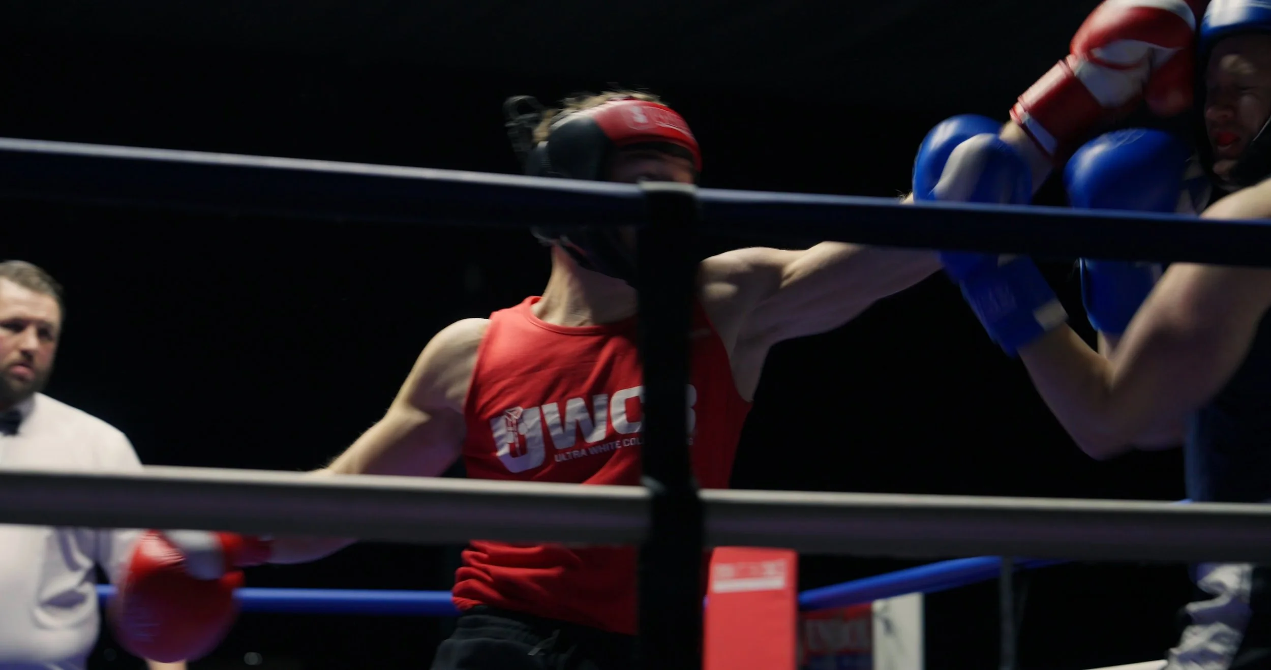 Boxer in red shorts and black headgear throws a punch at opponent in blue gloves during a match, with referee observing.