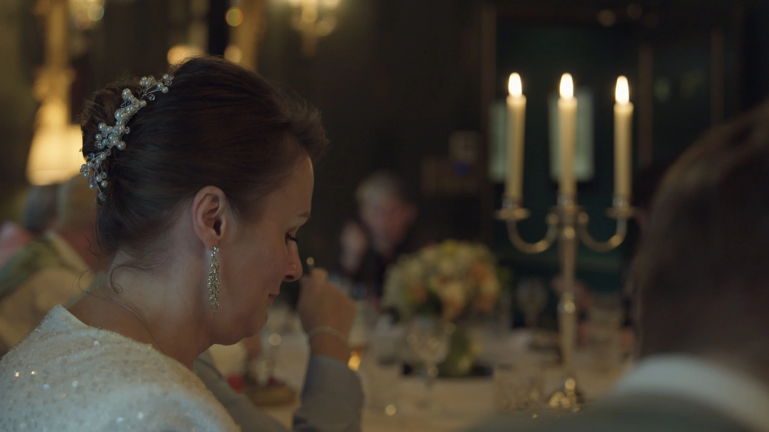 A woman with dark hair styled in an updo, wearing a pearl and crystal hair accessory and dangling earrings, sitting at a candlelit table during a formal event, with other guests blurred in the background.