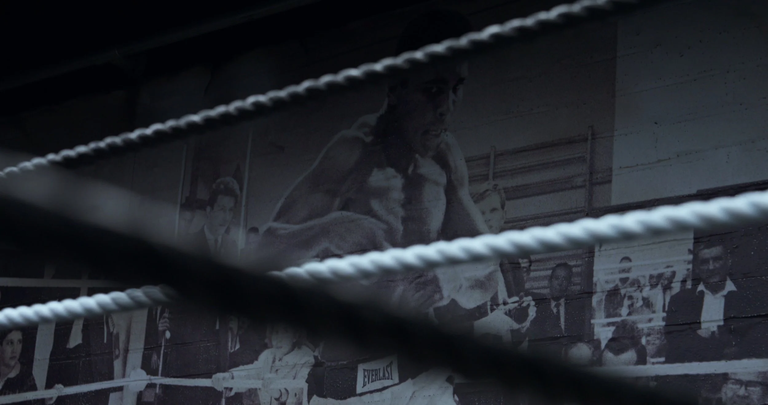 A black-and-white photograph of a boxing ring with a boxer sitting on the ropes, with a crowd and framed pictures in the background, viewed through the ropes of the ring.
