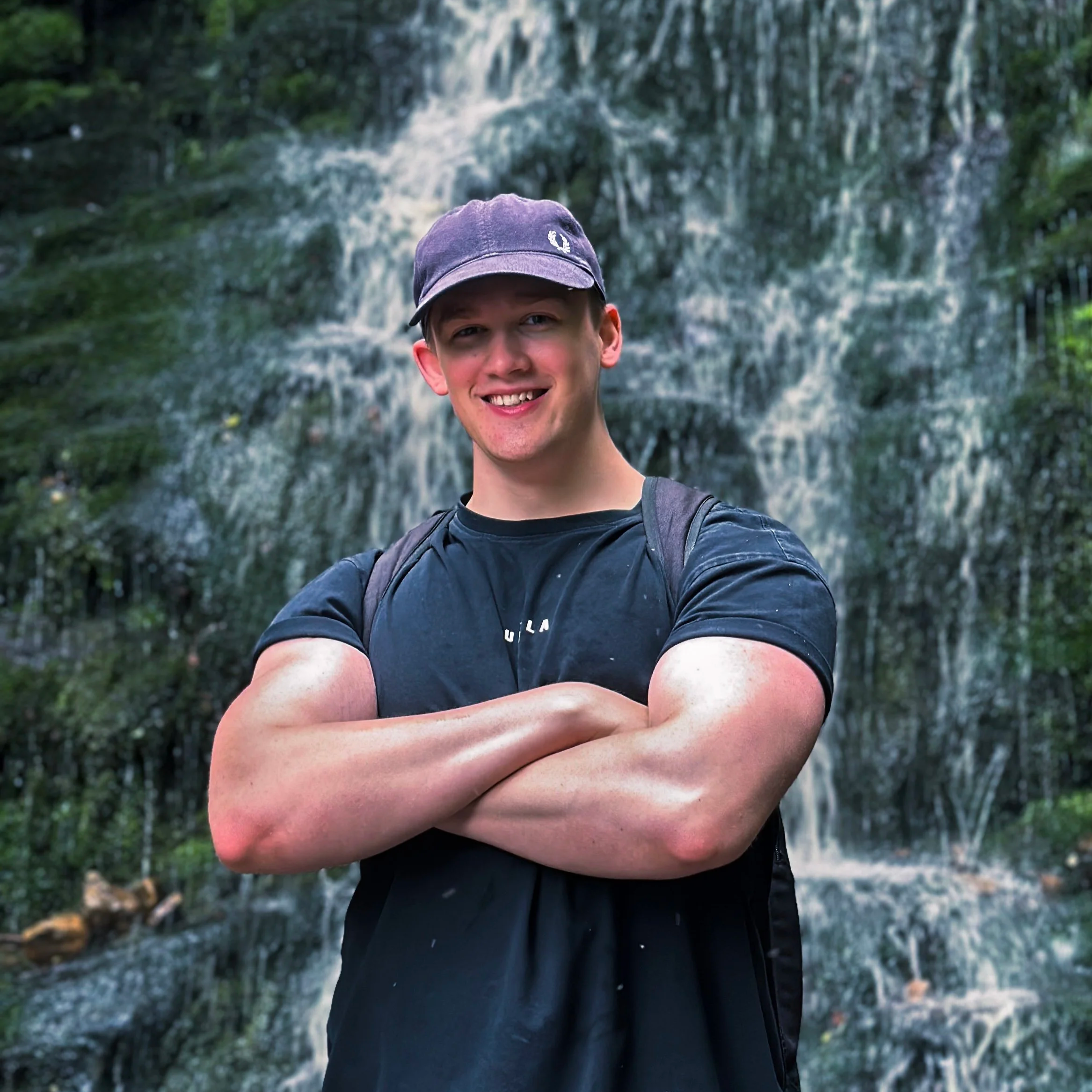 A young man with a baseball cap, black t-shirt, and backpack smiling with arms crossed in front of a waterfall in a forest setting.
