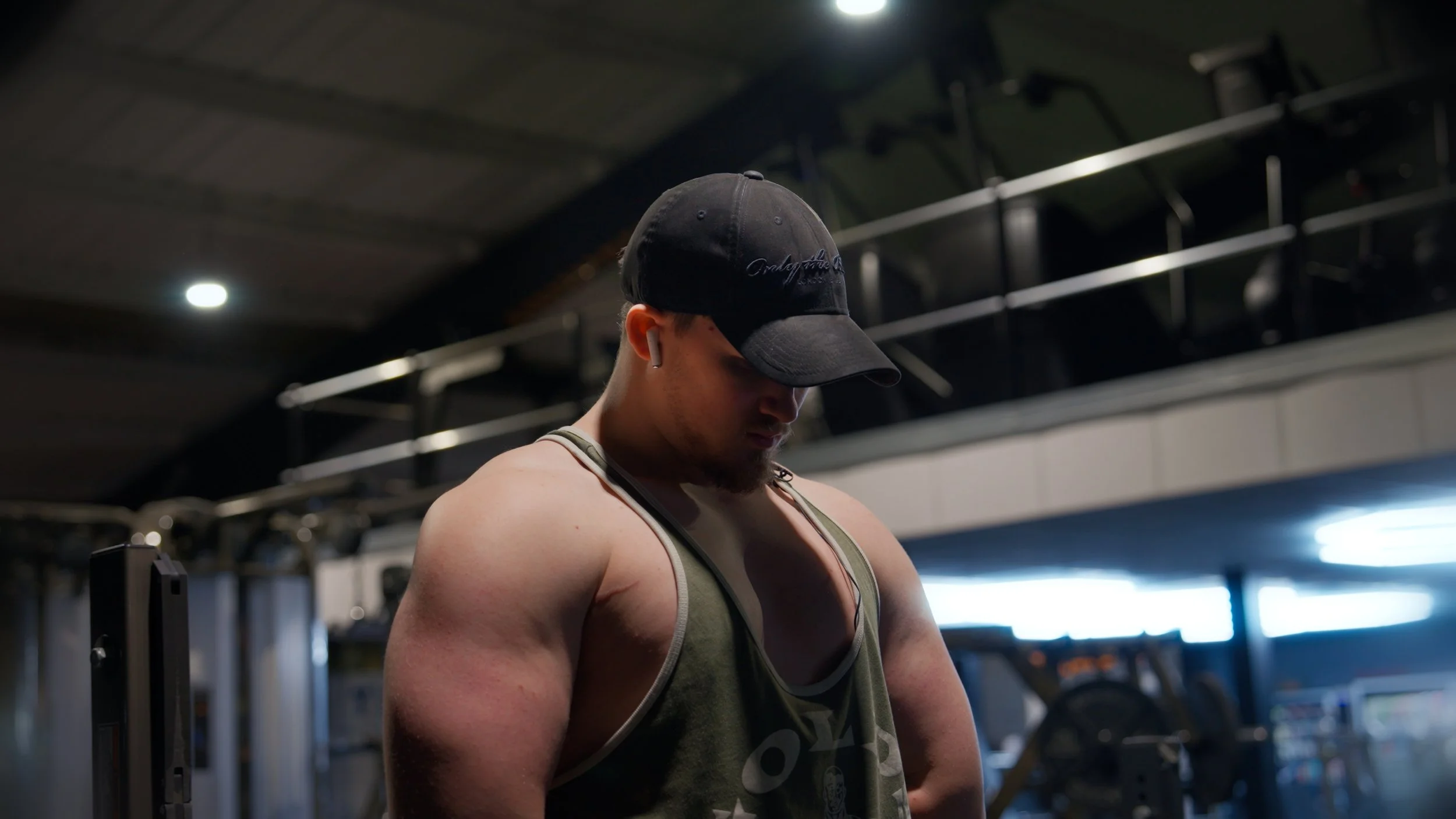 A muscular man wearing a black cap, green tank top, and wireless earbuds, looking down in a gym.