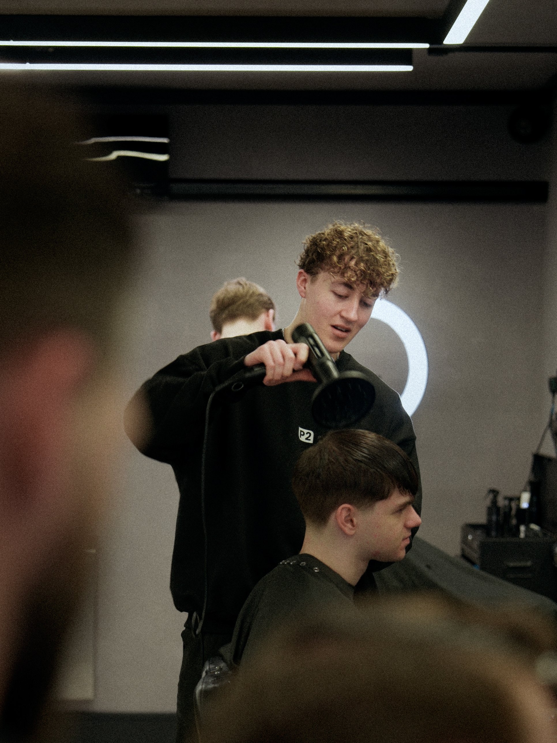 A young man with curly hair is blow-drying the haircut of another young man with straight hair at a hair salon.