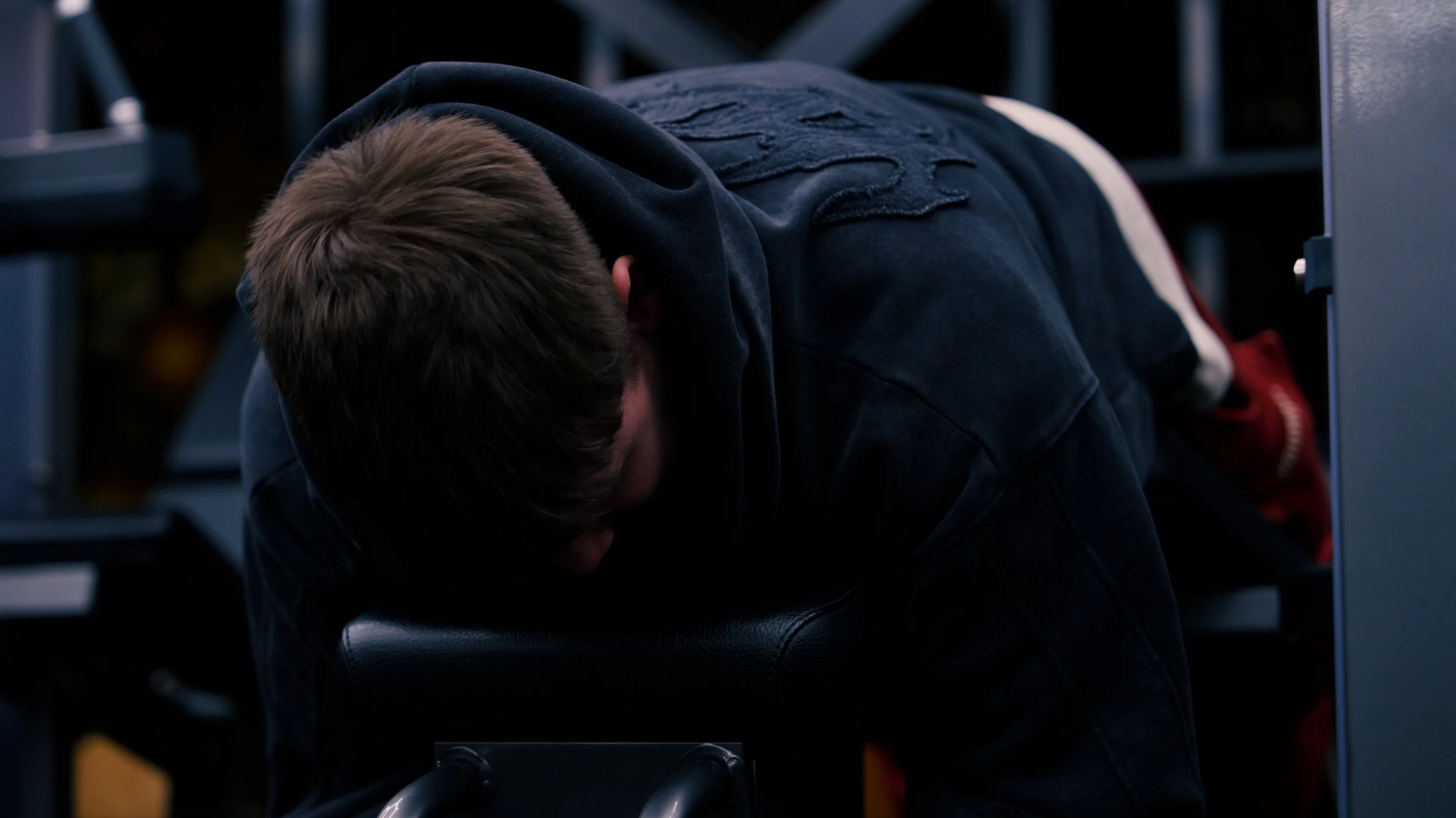 A person wearing a black hoodie and ripped jeans leaning on a black stool with their head down in a dimly lit room.