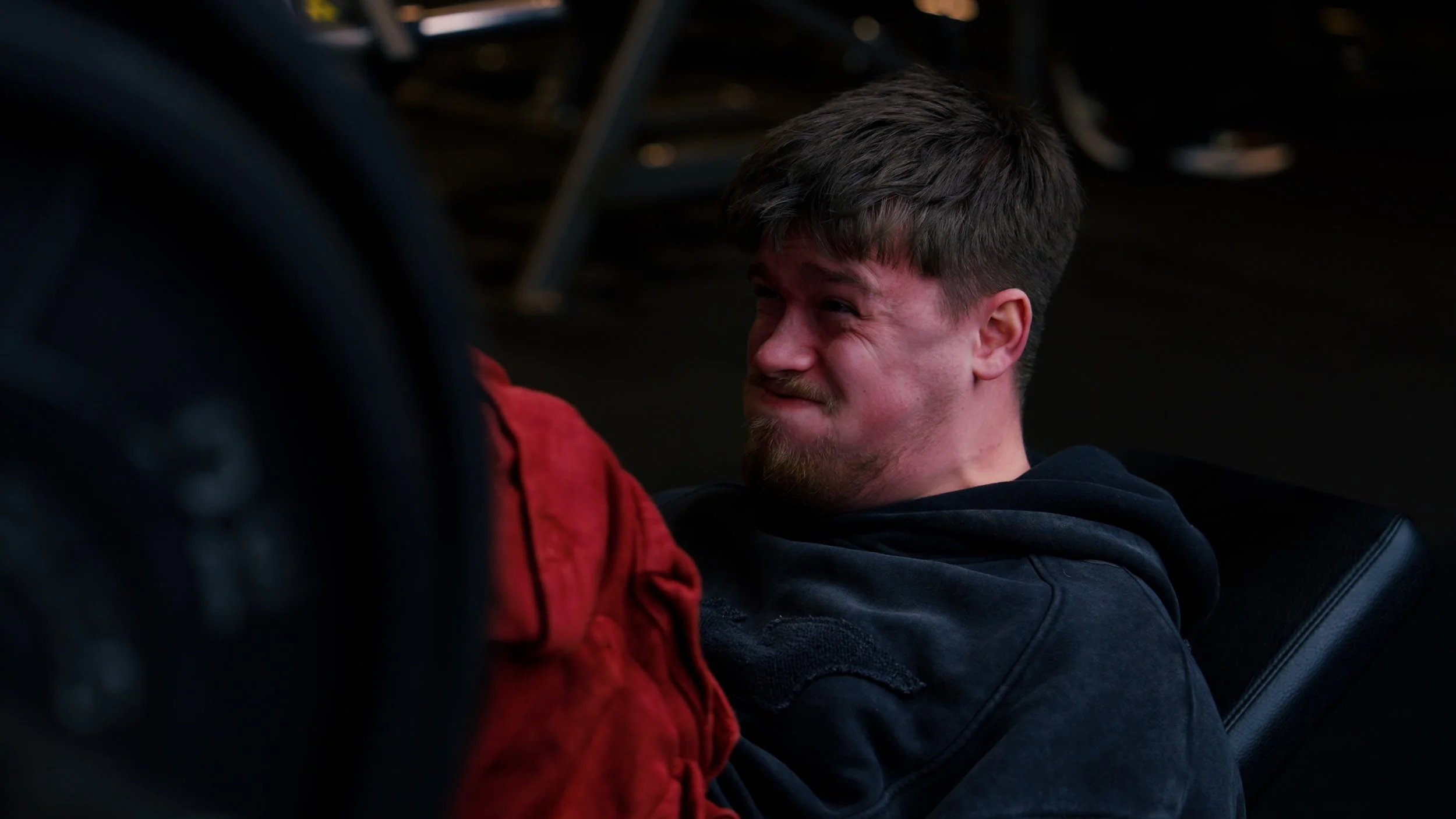 A man with short brown hair, a beard, and a mustache is sitting on a black chair. He is wearing a black hoodie and appears to be grimacing or showing discomfort.