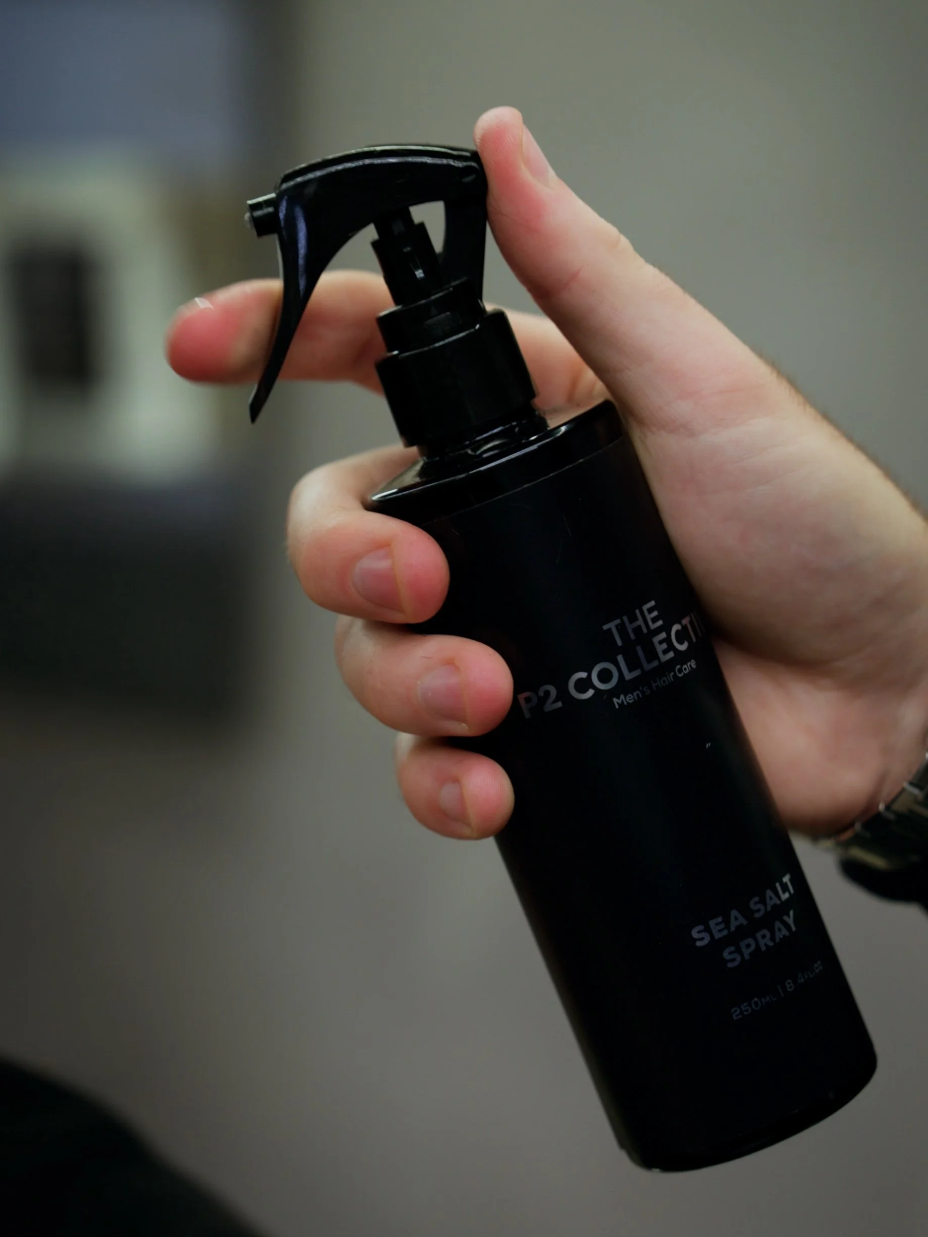Close-up of a person holding a black spray bottle labeled 'Sea Salt Spray' from 'The P2 Collection Men's Hair Care' in a gray background.