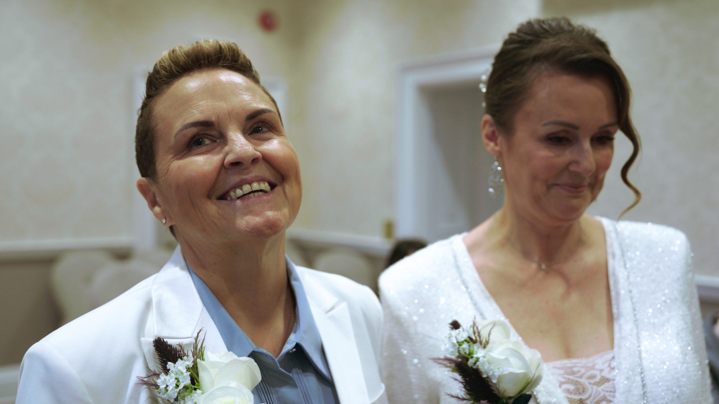 Two women dressed in formal attire with corsages, standing in an indoor setting, smiling and looking in different directions.