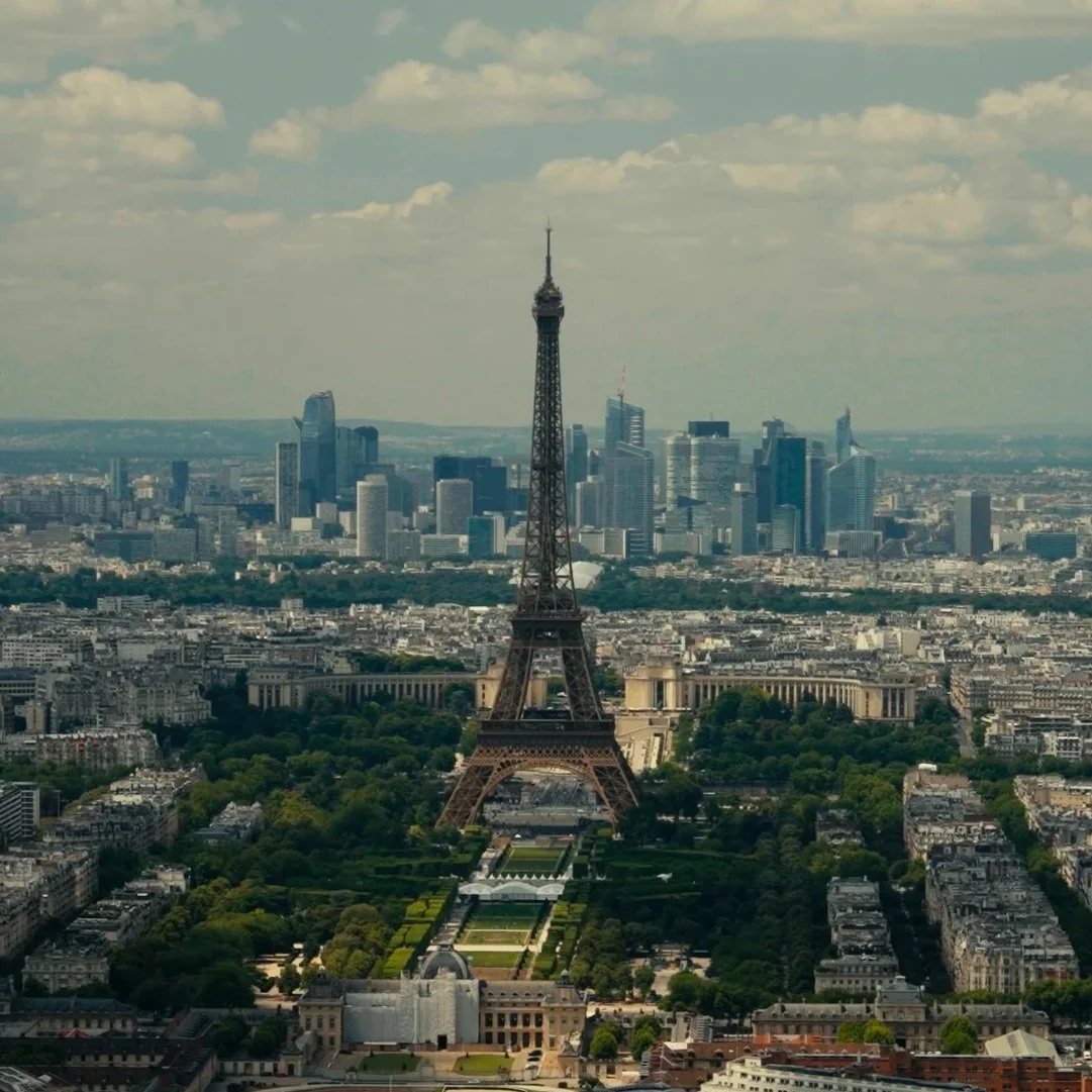 View of the Paris cityscape featuring the Eiffel Tower in the foreground and modern skyscrapers in the background, with a partly cloudy sky.