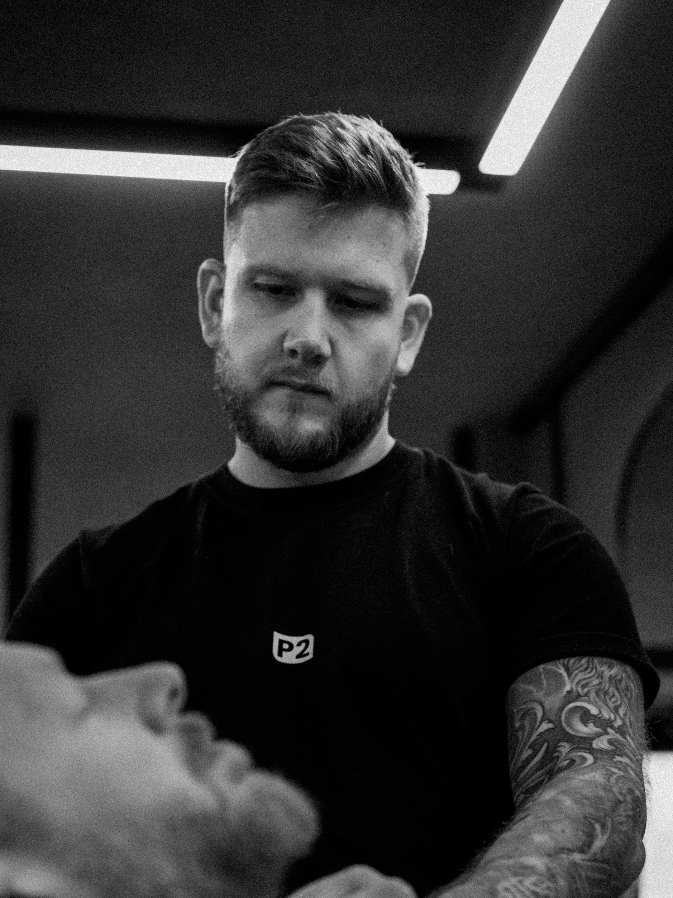 Black and white photo of a young man with a beard and short hair, wearing a black t-shirt with a small logo on the chest, looking down at something not visible in the image, in an indoor setting with ceiling lighting.