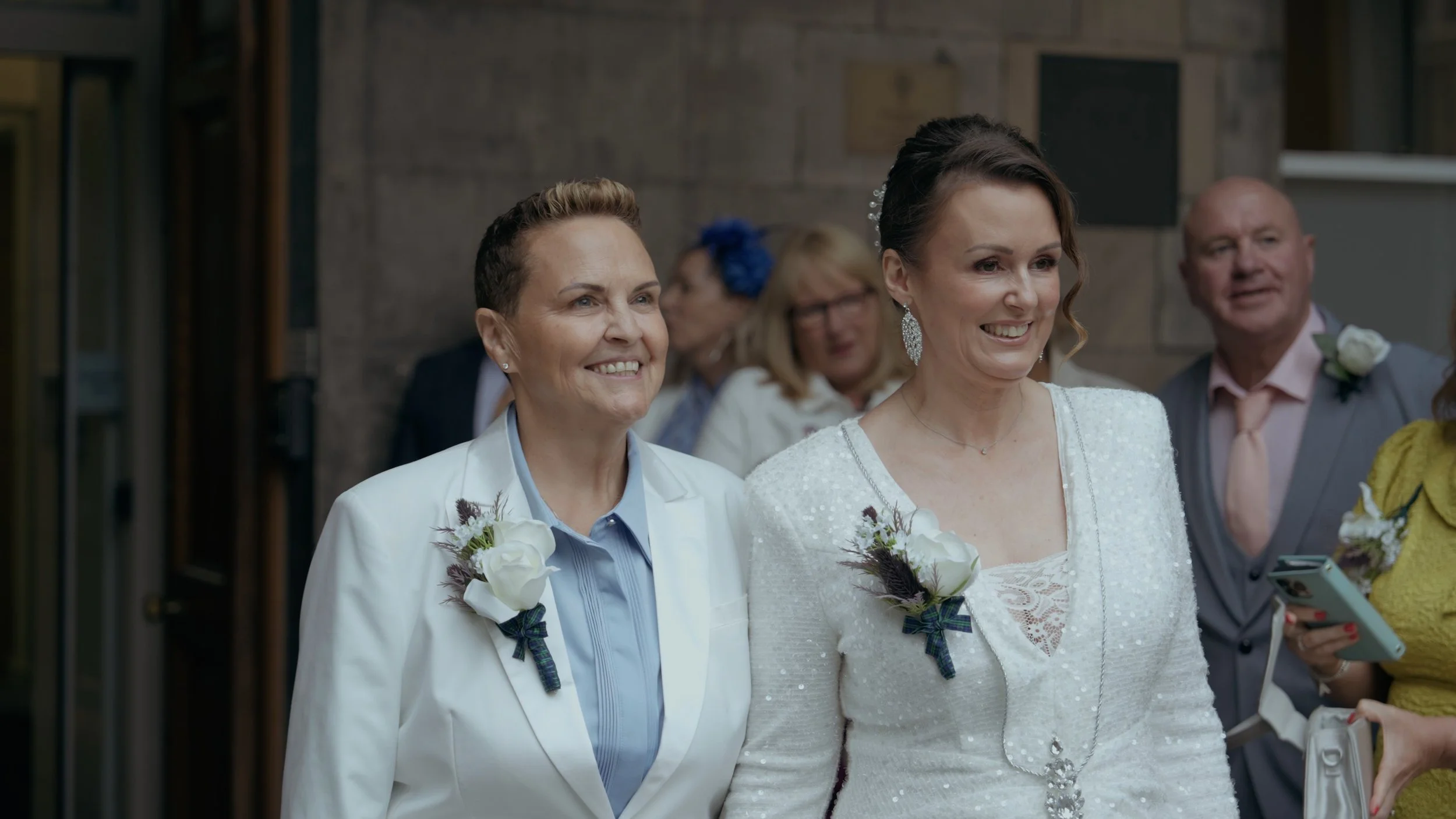 Two women smiling at a wedding, wearing white wedding attire with boutonnières, surrounded by guests.