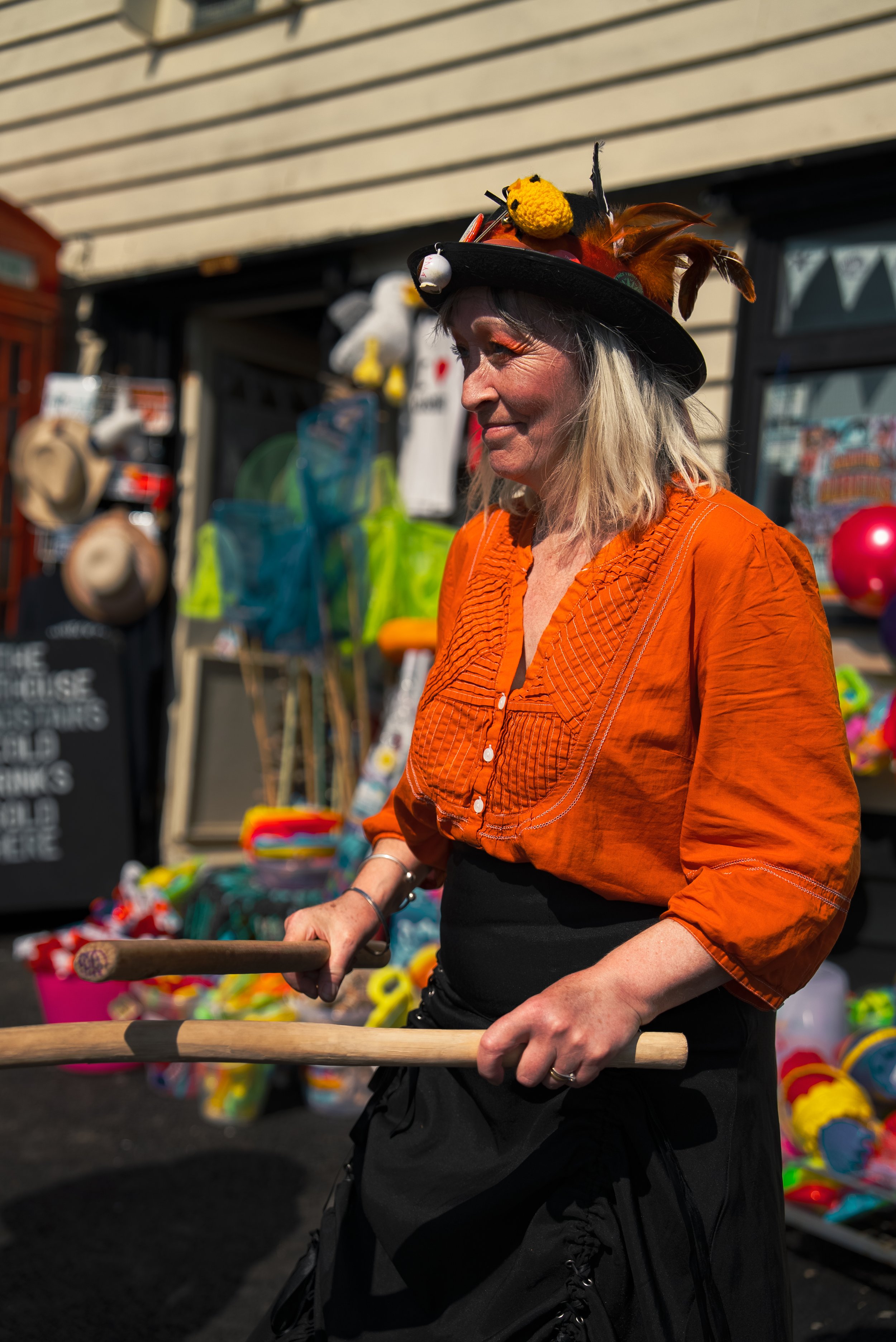 A woman wearing an elaborate hat decorated with feathers and plush toys, smiling as she walks through an outdoor market, holding a wooden stick.