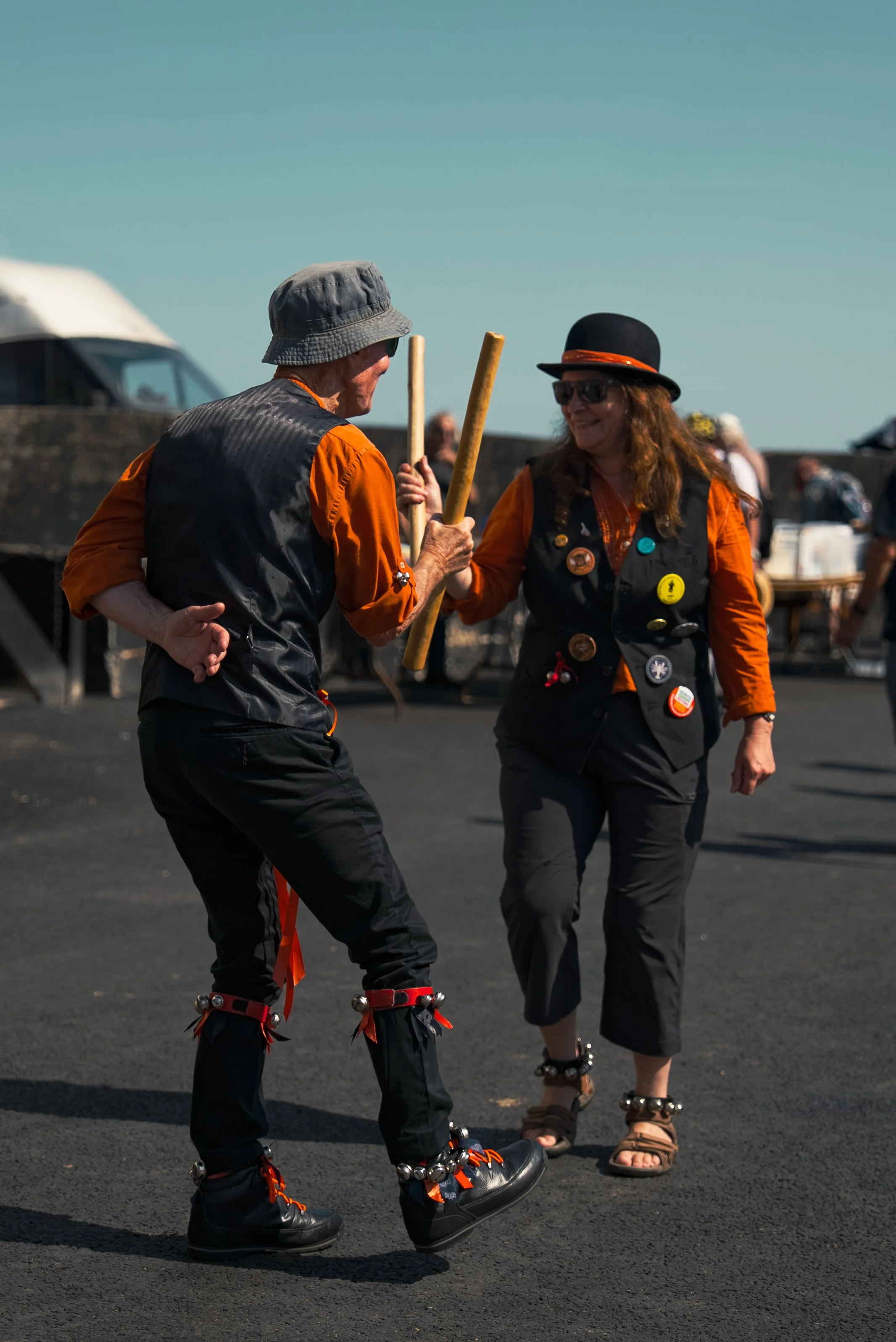 Two people dancing outdoors on a sunny day, both wearing black pants and orange shirts with black vests decorated with buttons and pins. They are holding sticks and wearing jingle bells around their ankles.