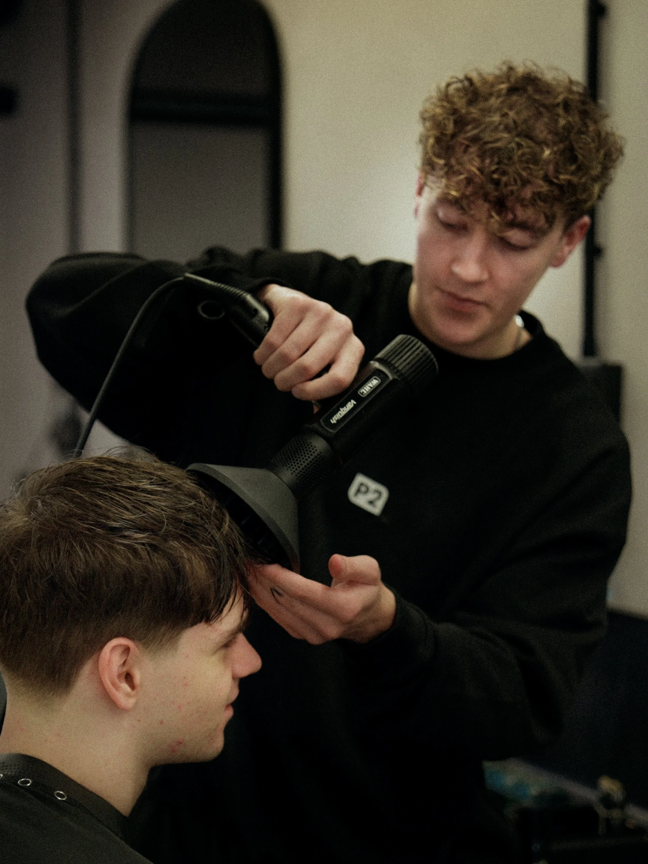 A hairstylist using a professional hairdryer on a young man's hair in a salon.