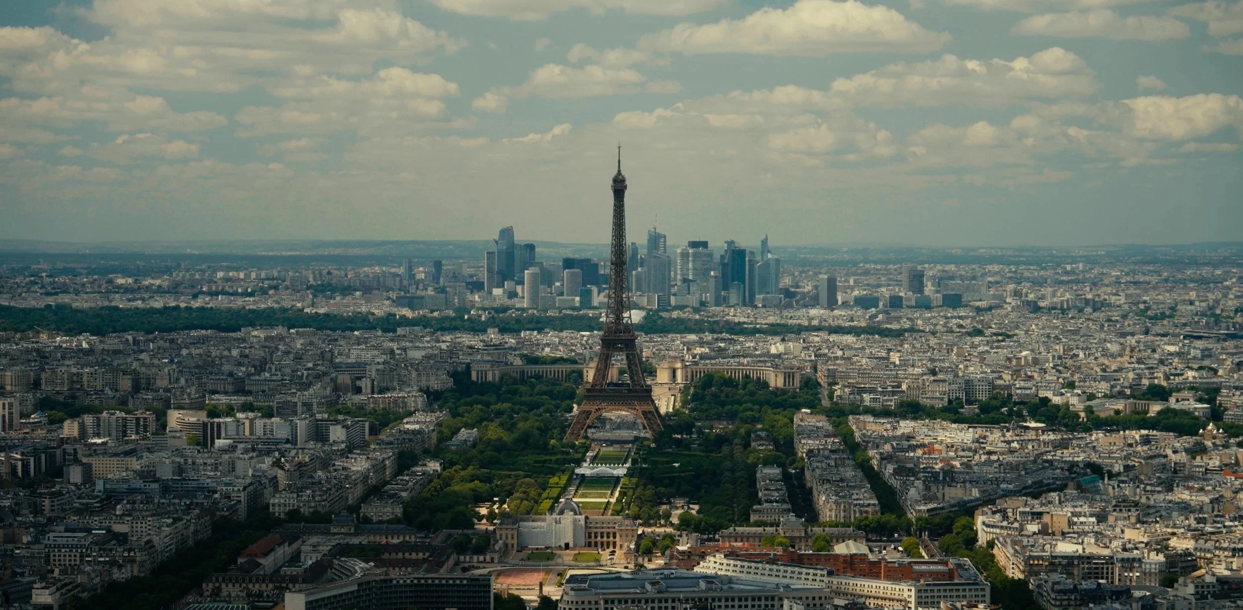 Aerial view of Paris featuring the Eiffel Tower in the foreground and the La Défense business district with skyscrapers in the background under partly cloudy skies.