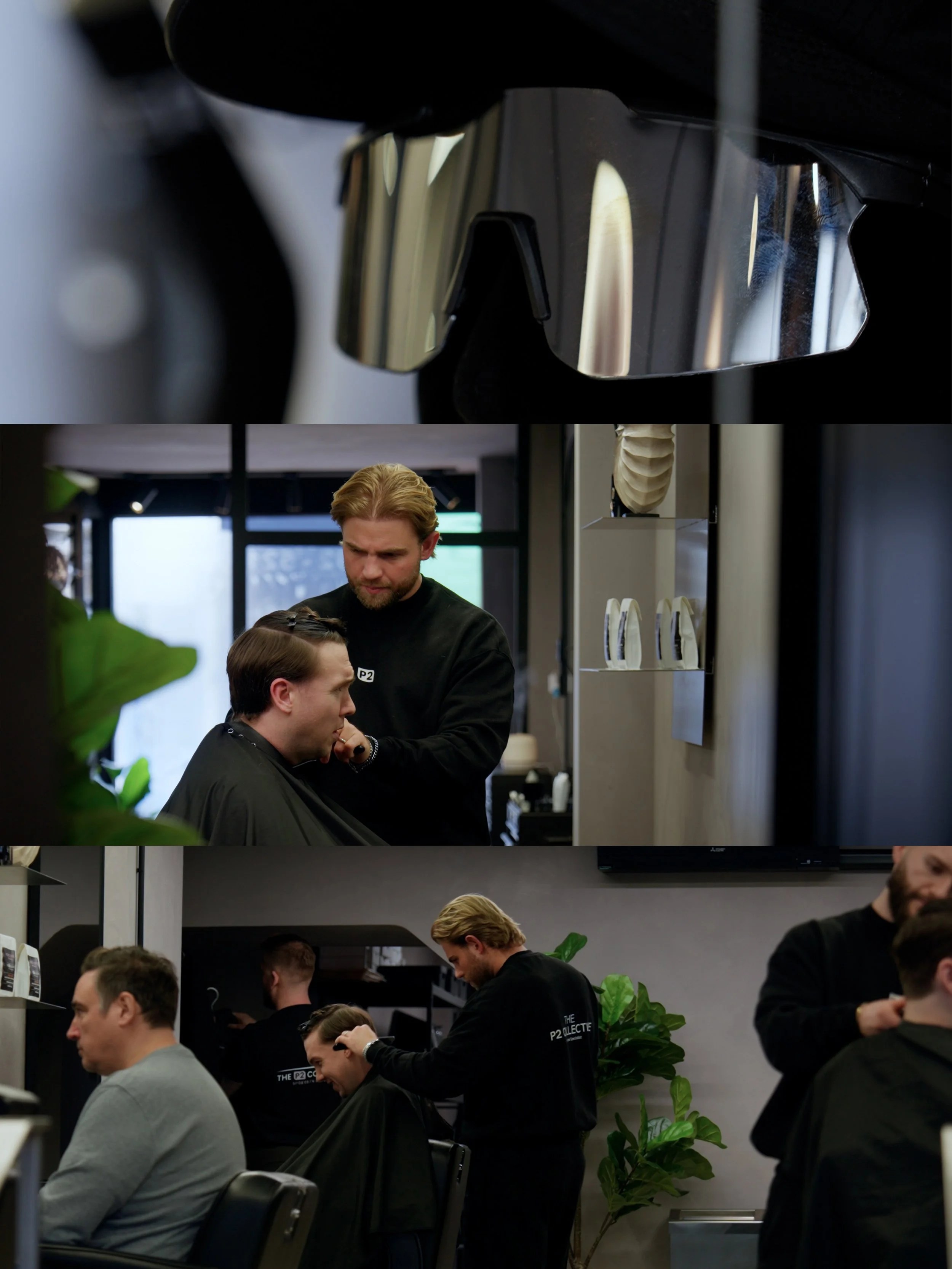 A man in a black salon cape getting a haircut by a hairstylist with blond hair in a salon. Other clients and stylists are visible in the background, with salon equipment and plants, in a modern salon setting.