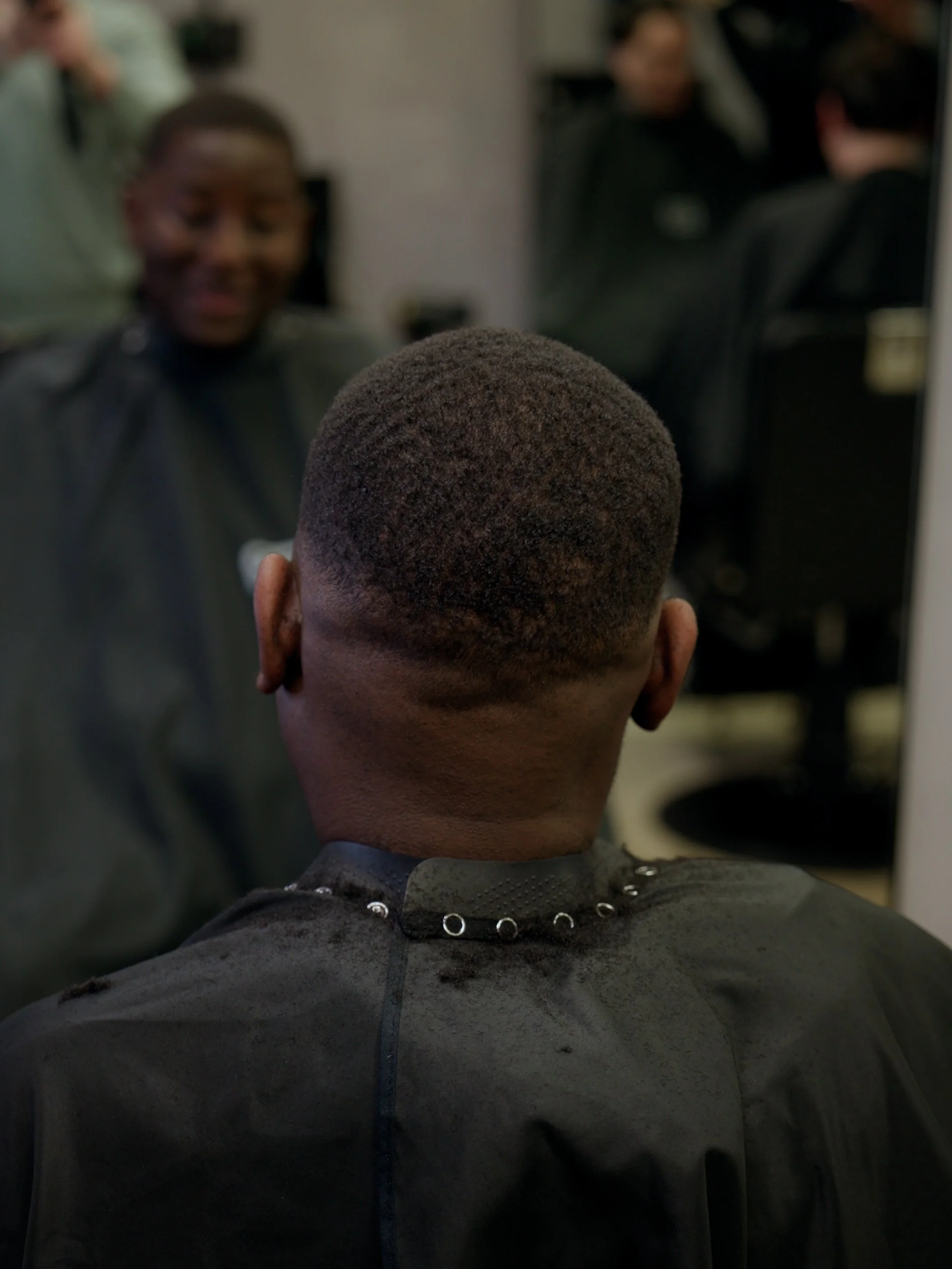 Man with a fresh haircut sitting in front of a mirror at a barber shop, barber smiling in the background.